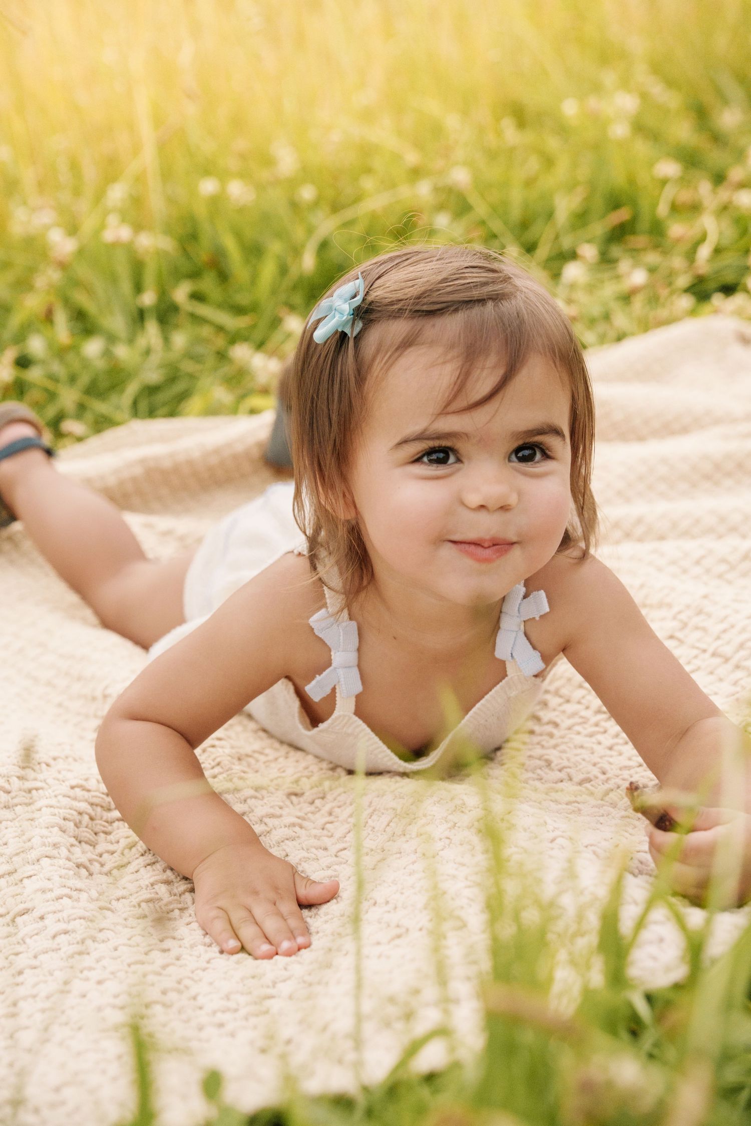 Adorable baby girl with hair clip lying on blanket in sunny green grass field.