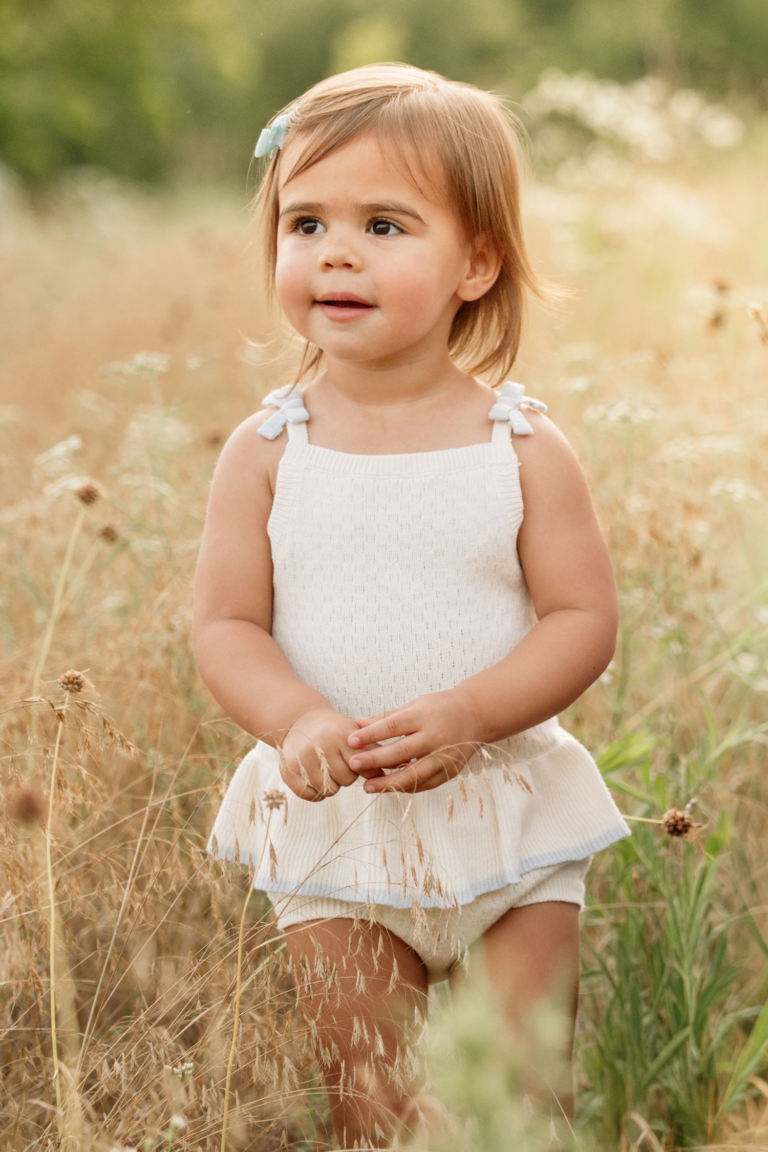 Toddler girl in white romper standing in golden meadow with wildflowers, looking up curiously on sunny day.
