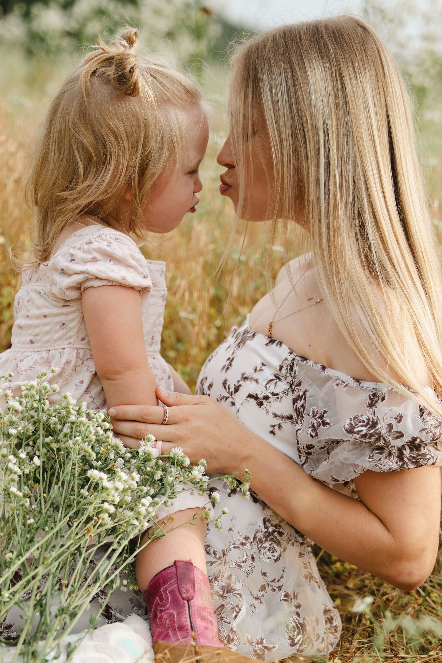 A blonde mother and toddler daughter in matching floral dresses share a tender nose-to-nose moment outdoors.