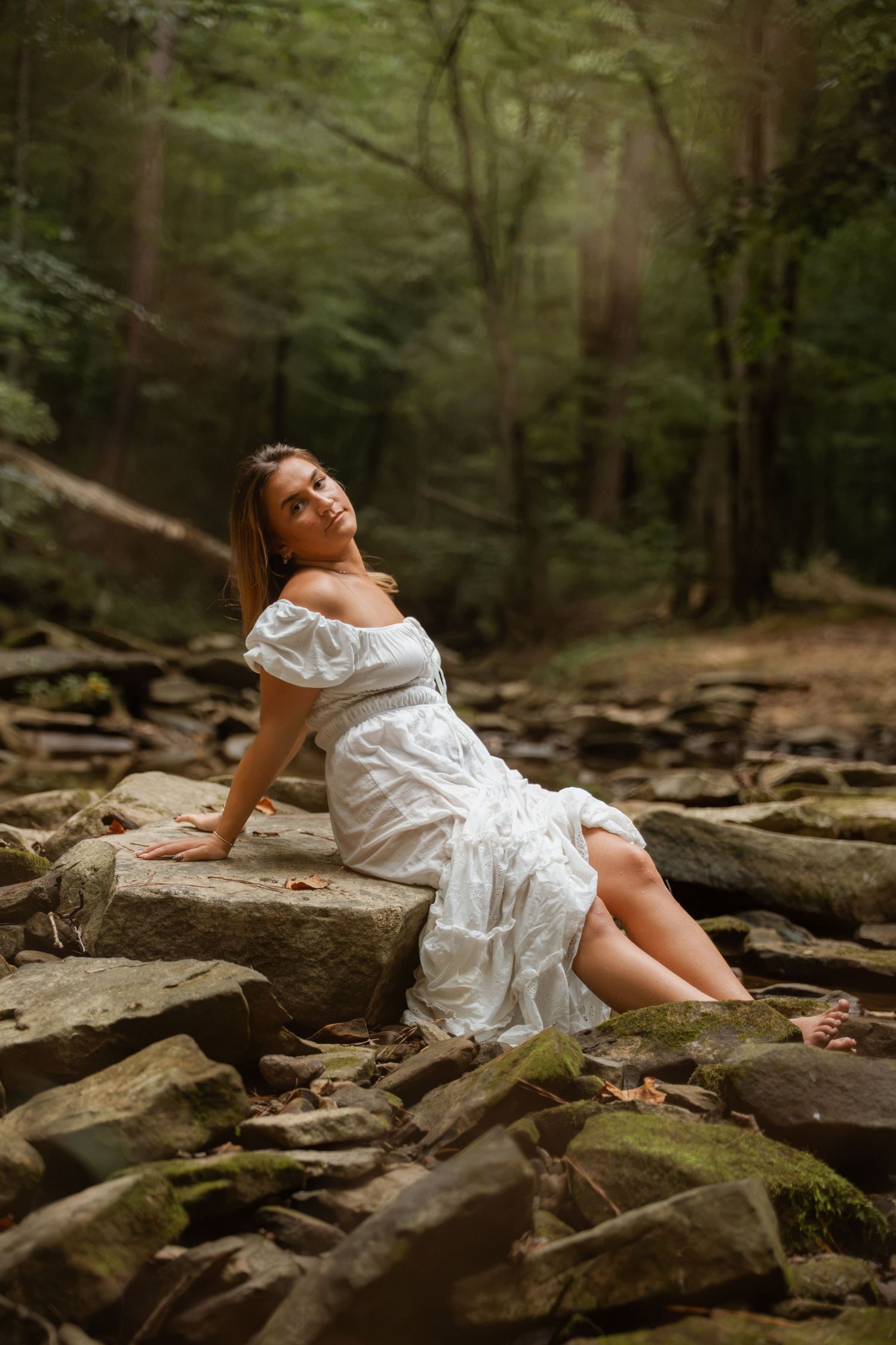 Smiling woman in white off-shoulder dress sitting on mossy rocks in a lush green forest setting.