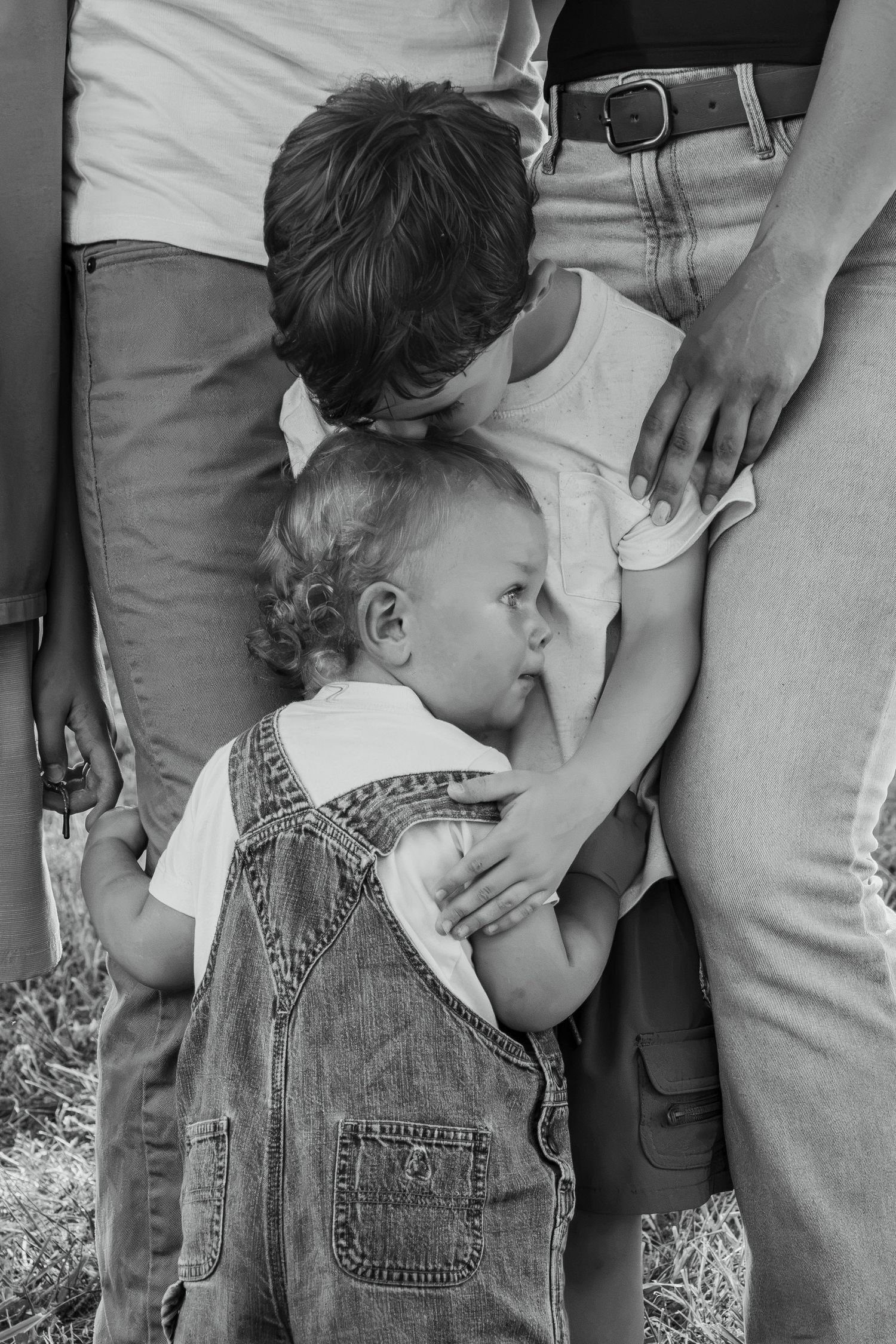 Black and white photo of two young children clinging to adults legs outdoors, toddler in denim overalls.