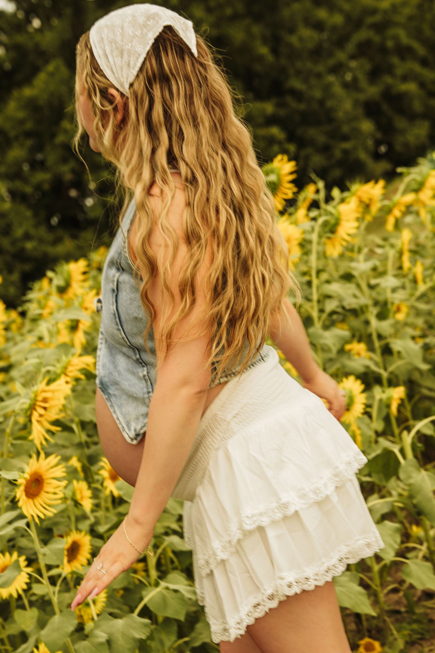Young woman with wavy blonde hair wearing denim top and white ruffled skirt posing in a sunflower field.
