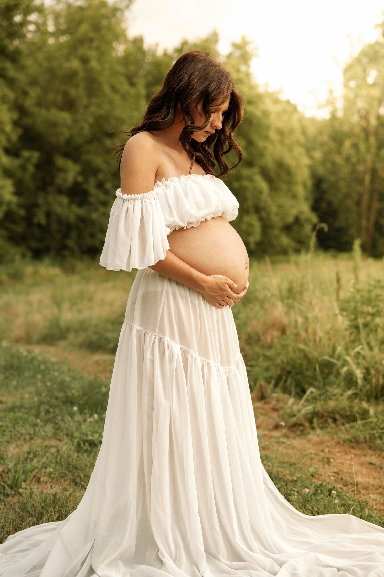 Pregnant woman in white off-shoulder gown cradling baby bump outdoors in golden hour light.