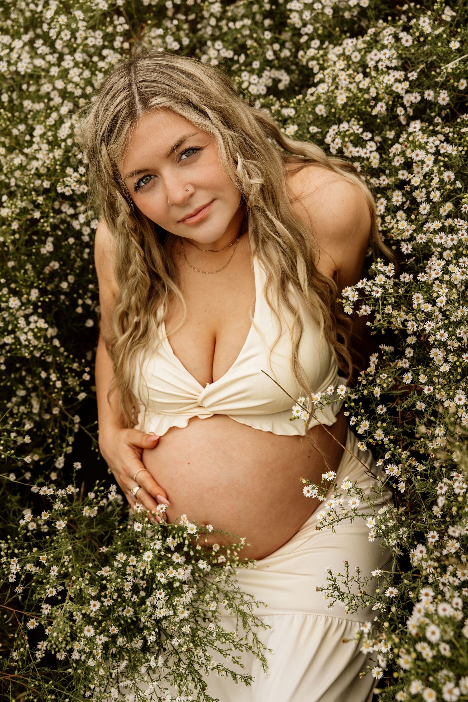 Pregnant woman in white crop top poses among blooming white wildflowers, cradling her baby bump outdoors.