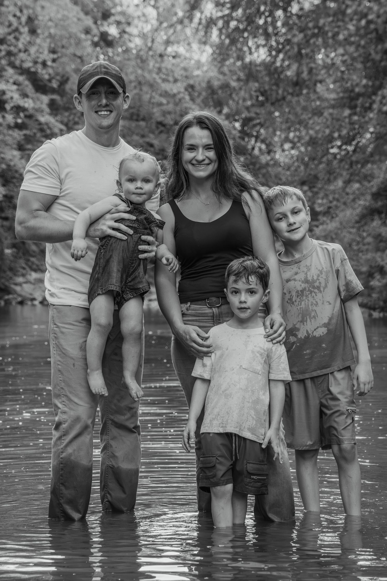 Black and white family portrait of parents with three young boys standing together in a shallow creek surrounded by trees.