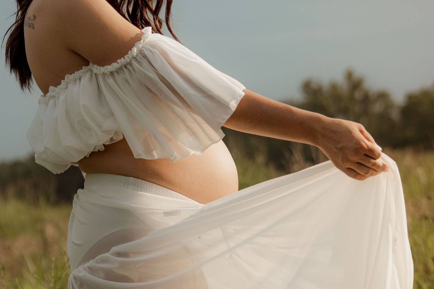 Pregnant woman in white off-shoulder crop top and flowing skirt holding fabric outdoors in a field.