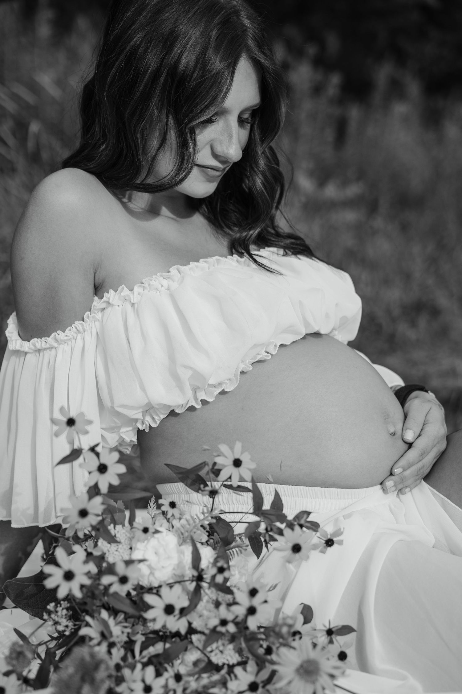 Black and white photo of pregnant woman in white dress cradling baby bump with flowers in foreground.