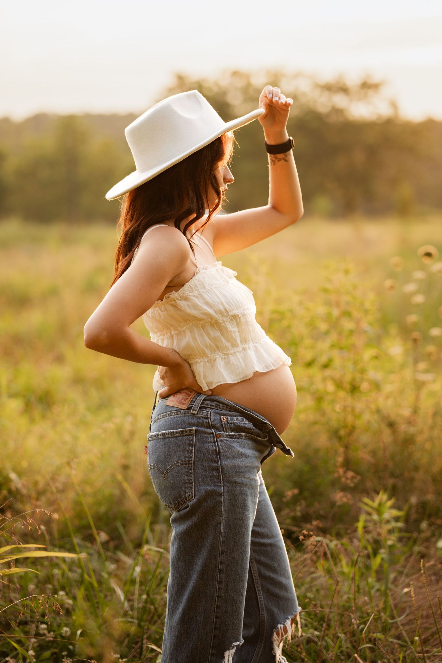 Pregnant woman in white hat and crop top posing in a glowing outdoor meadow.