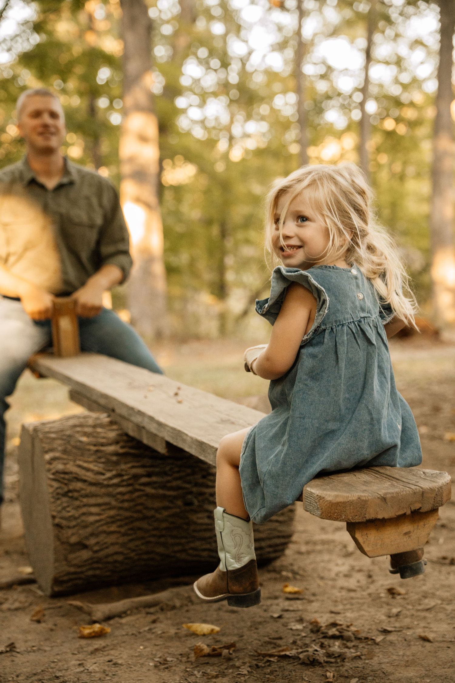 A smiling blonde girl in a teal dress and cowboy boots sits on a wooden seesaw with a man in a sunlit forest.