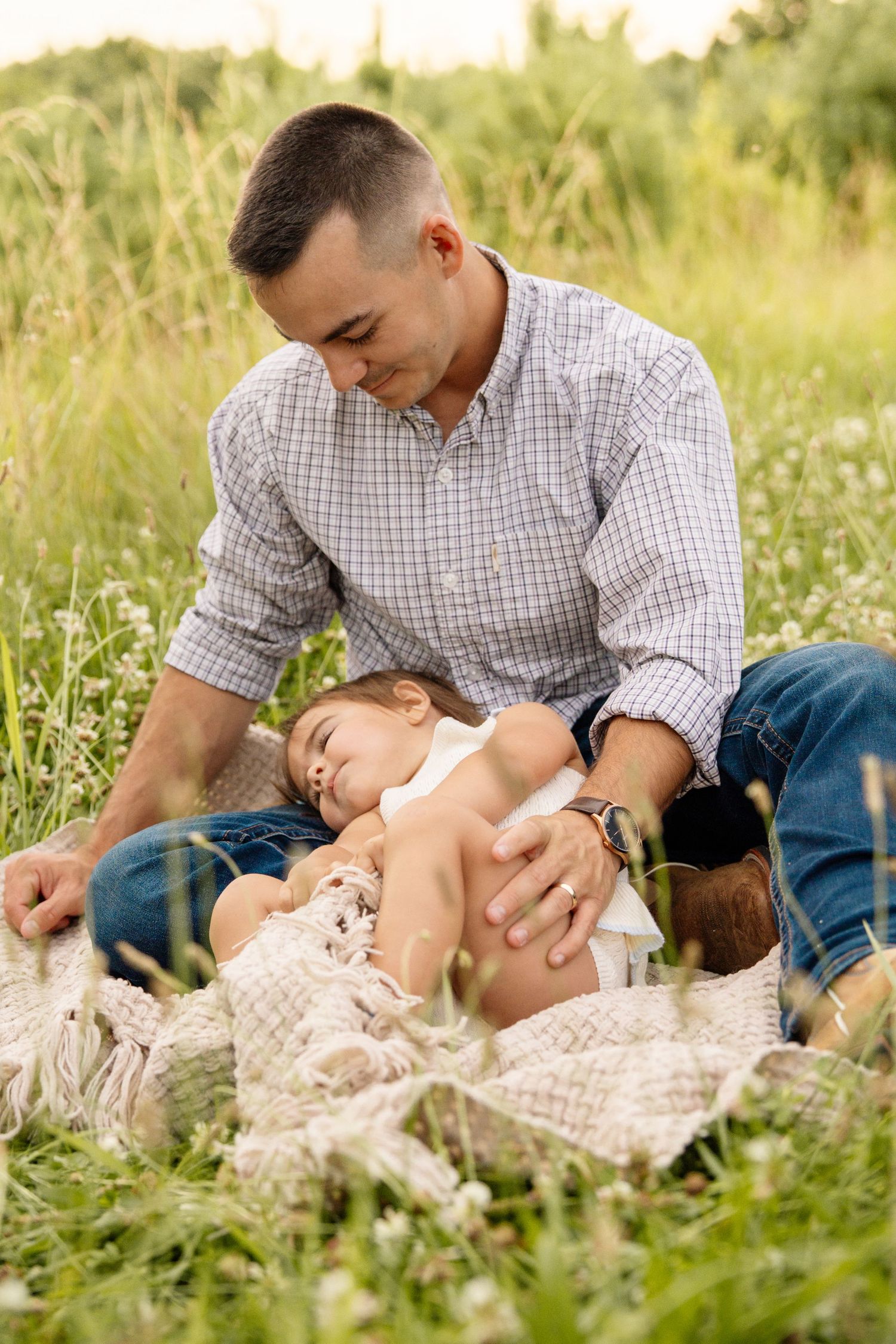 Father sitting in a grassy field cradling his sleepy toddler daughter on his lap outdoors.
