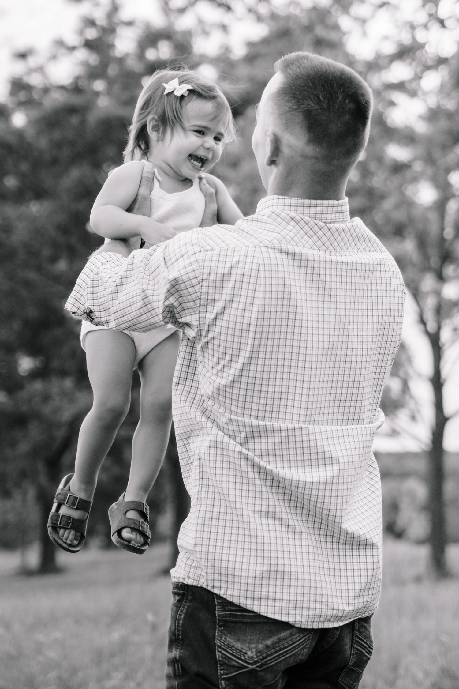 Father lifting laughing toddler girl in black and white outdoor park photo.