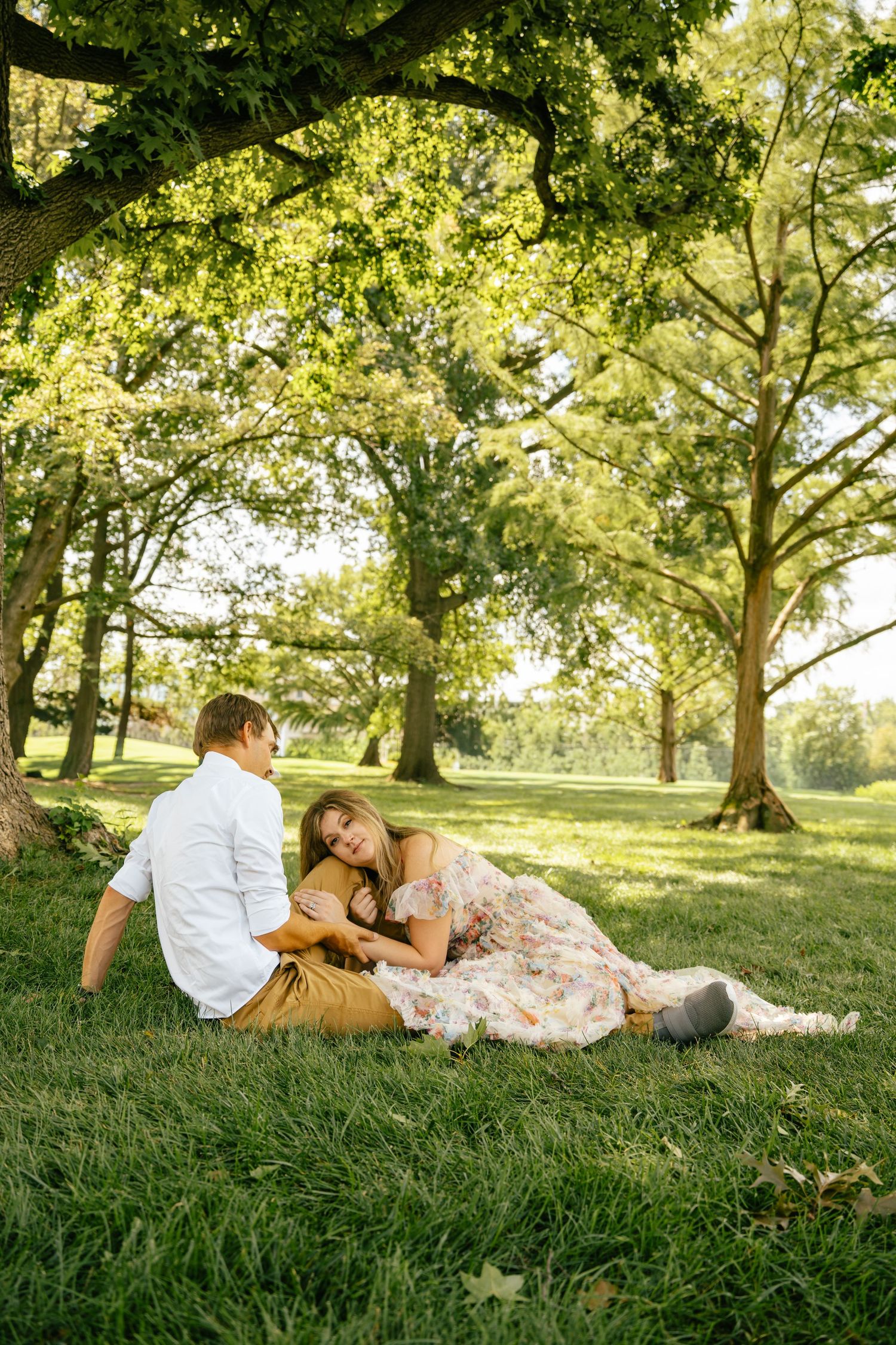 Couple relaxing on lush green grass under large trees in a sunlit park, woman in floral dress lying on mans lap.