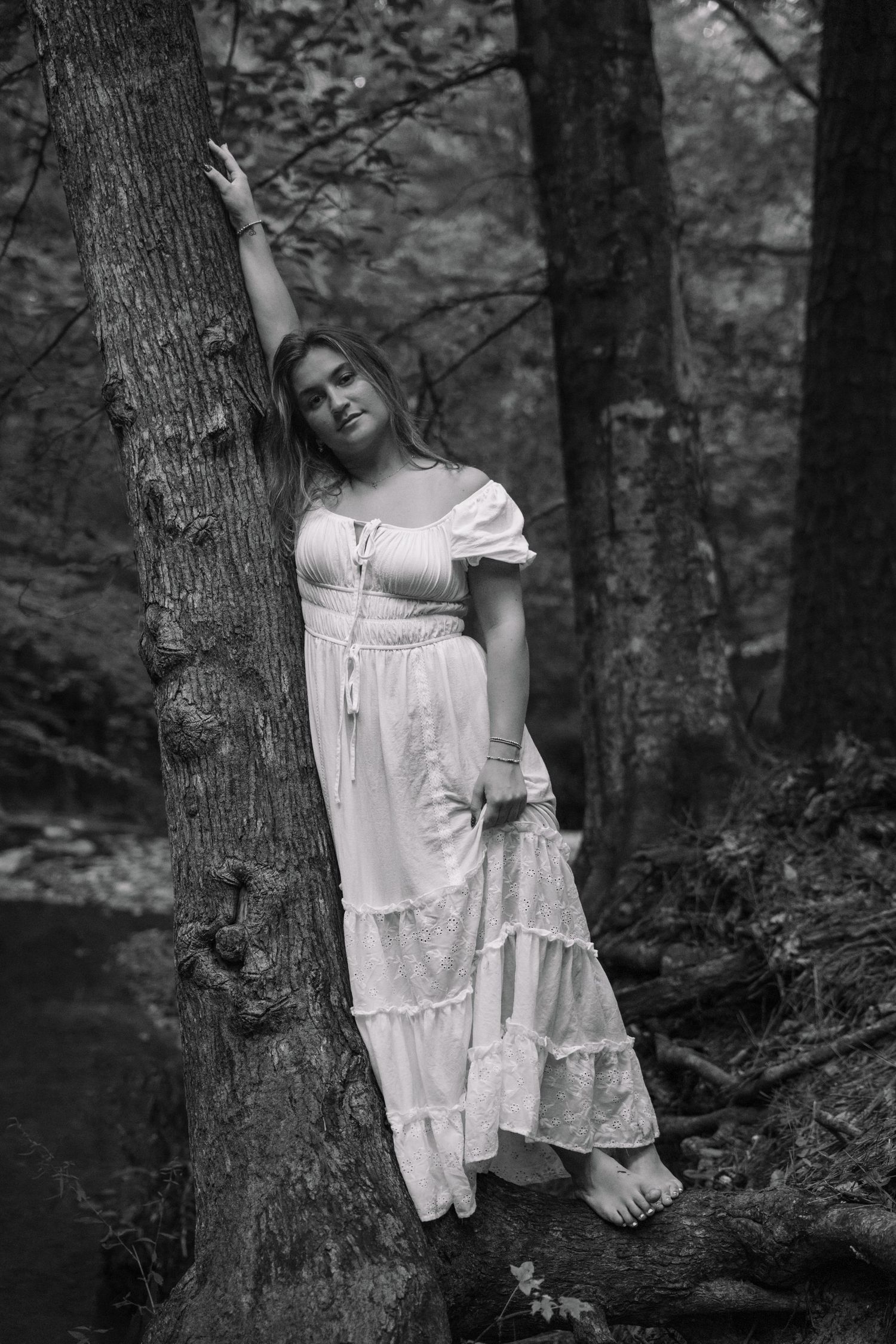 Black and white photo of a woman in white dress leaning against a forest tree.