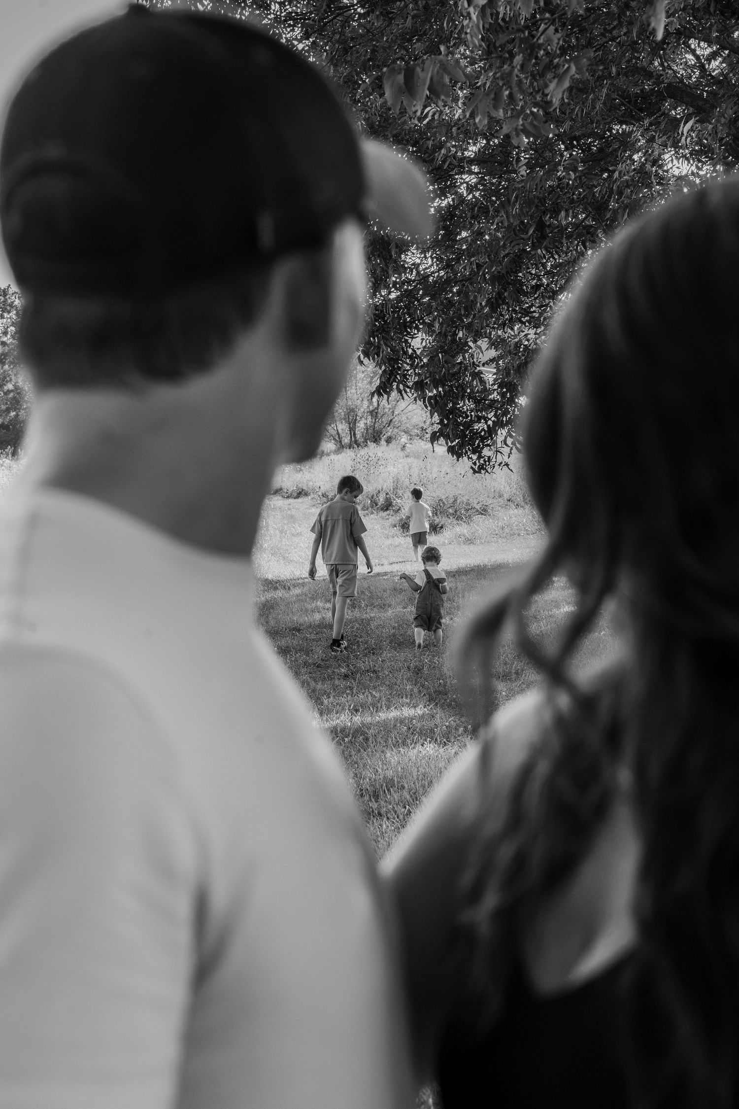 Black and white photo of parents watching their children play in a grassy park under a large tree.