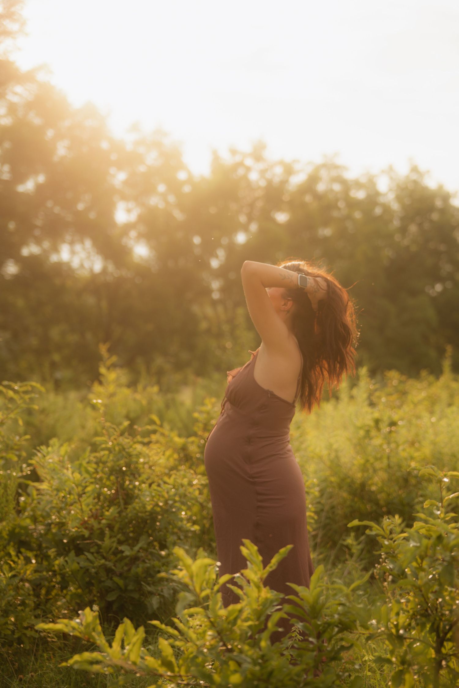 Pregnant woman in brown maxi dress turns away with hand in hair surrounded by green plants at sunset.