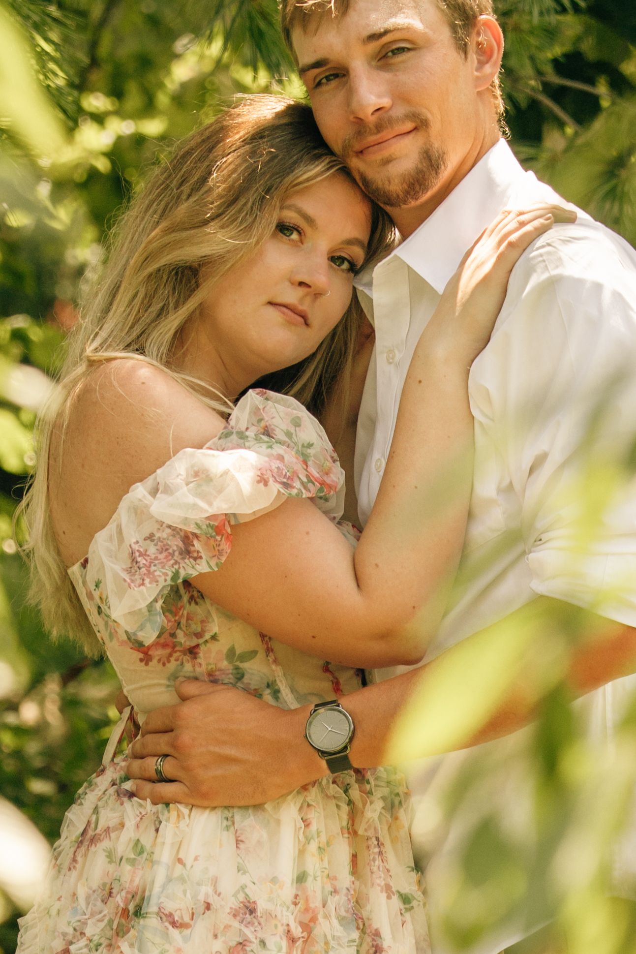 Couple embracing tenderly among green leaves, woman in floral dress leaning into mans chest.