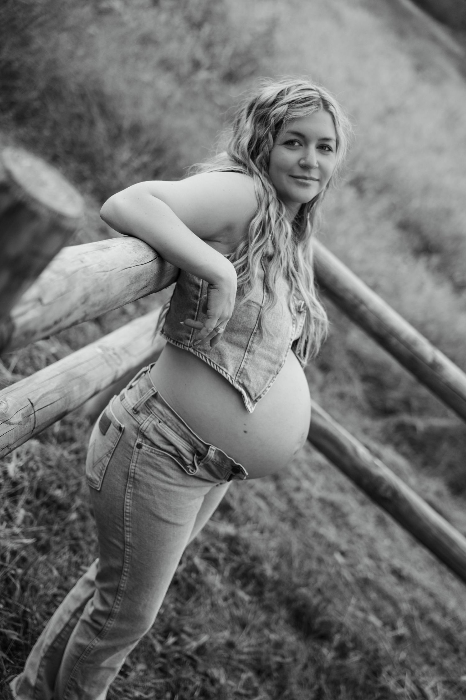 Black and white photo of pregnant woman leaning on wooden fence in field, wearing denim vest and jeans.