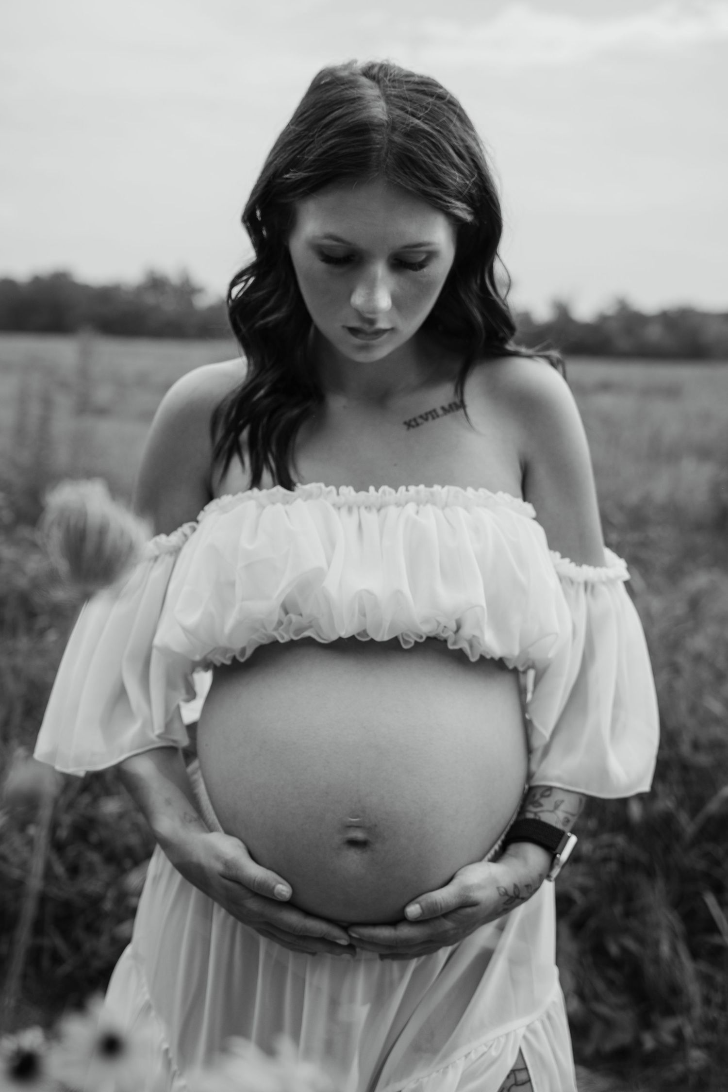 Black and white maternity photo of a pregnant woman in a white off-shoulder dress cradling her baby bump in a field.