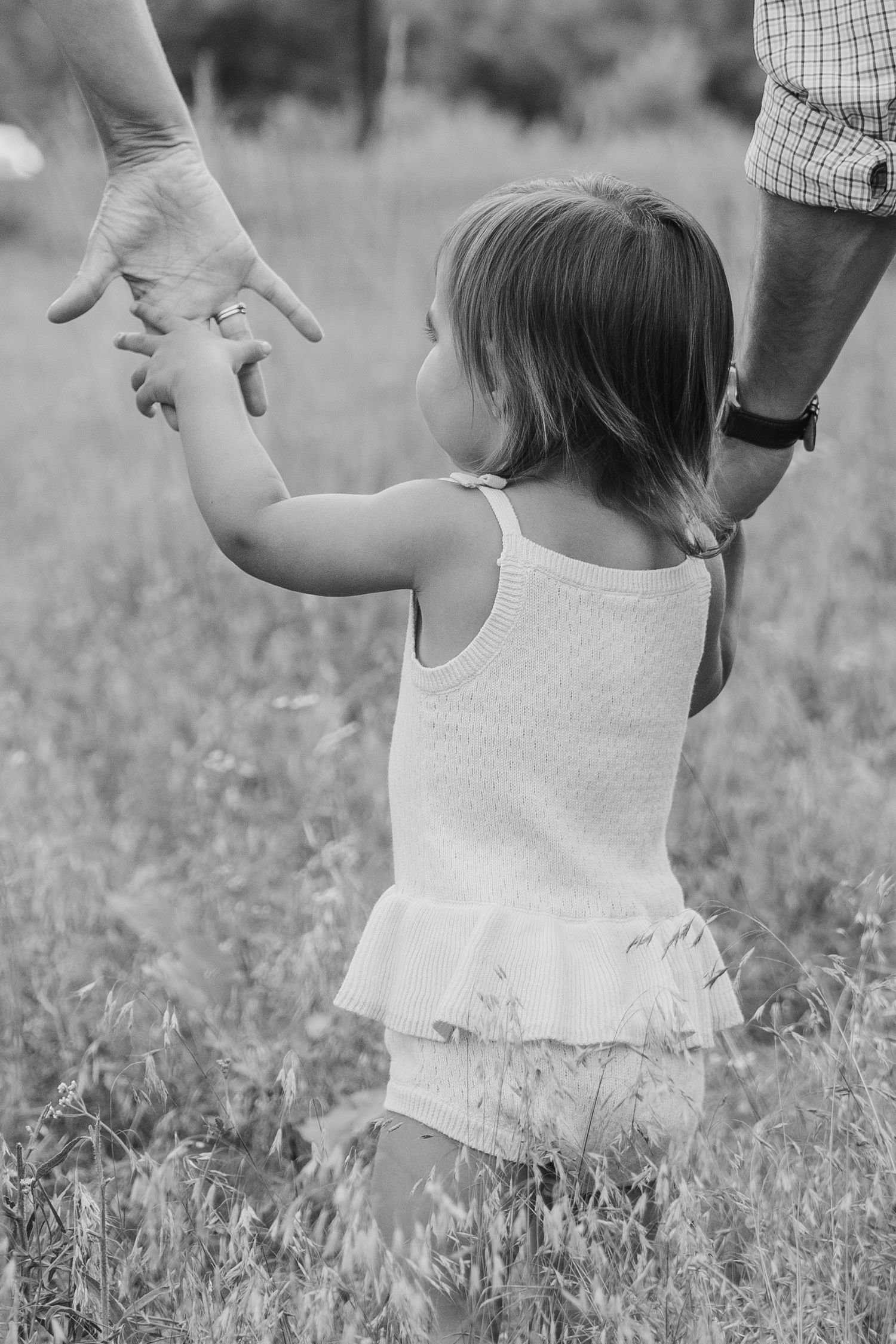 Black and white photo of a toddler girl in a white dress reaching up to hold her parent's hand in a grassy field.