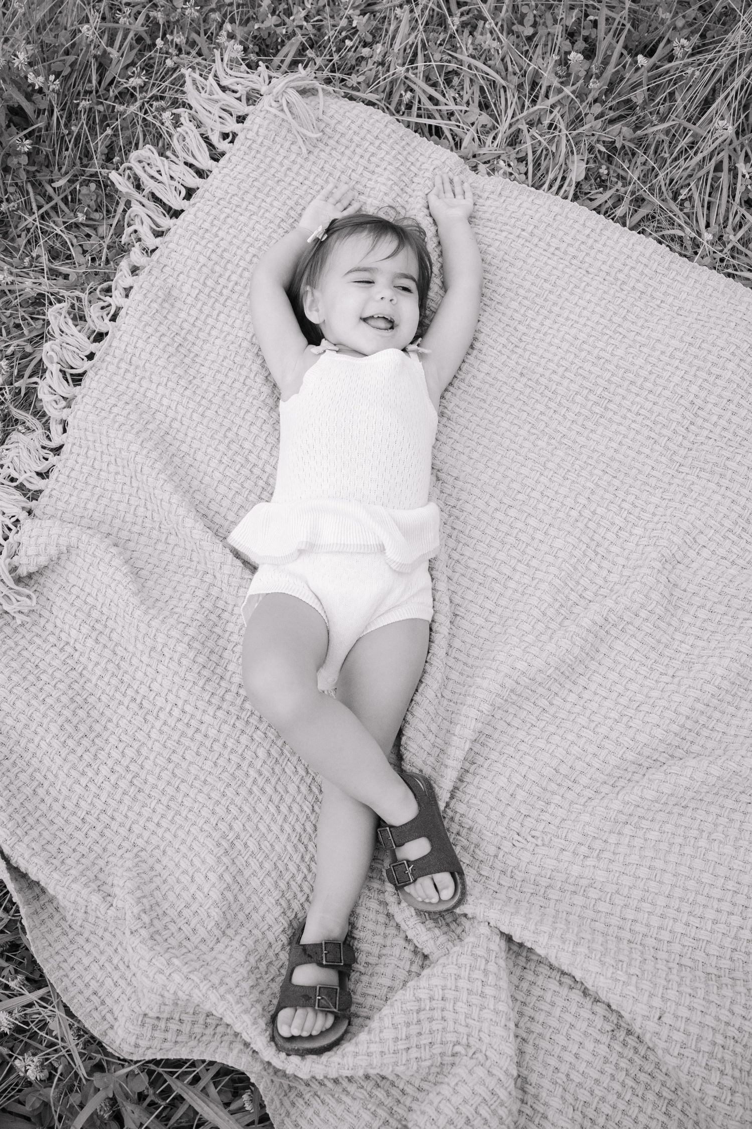 Black and white photo of a happy toddler lying on a blanket outdoors with arms raised and wearing sandals.
