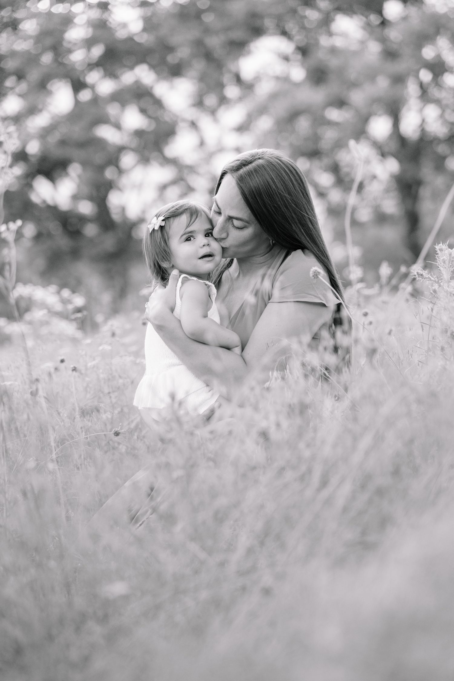 Black and white photo of mother tenderly kissing toddler daughter in wildflower field.