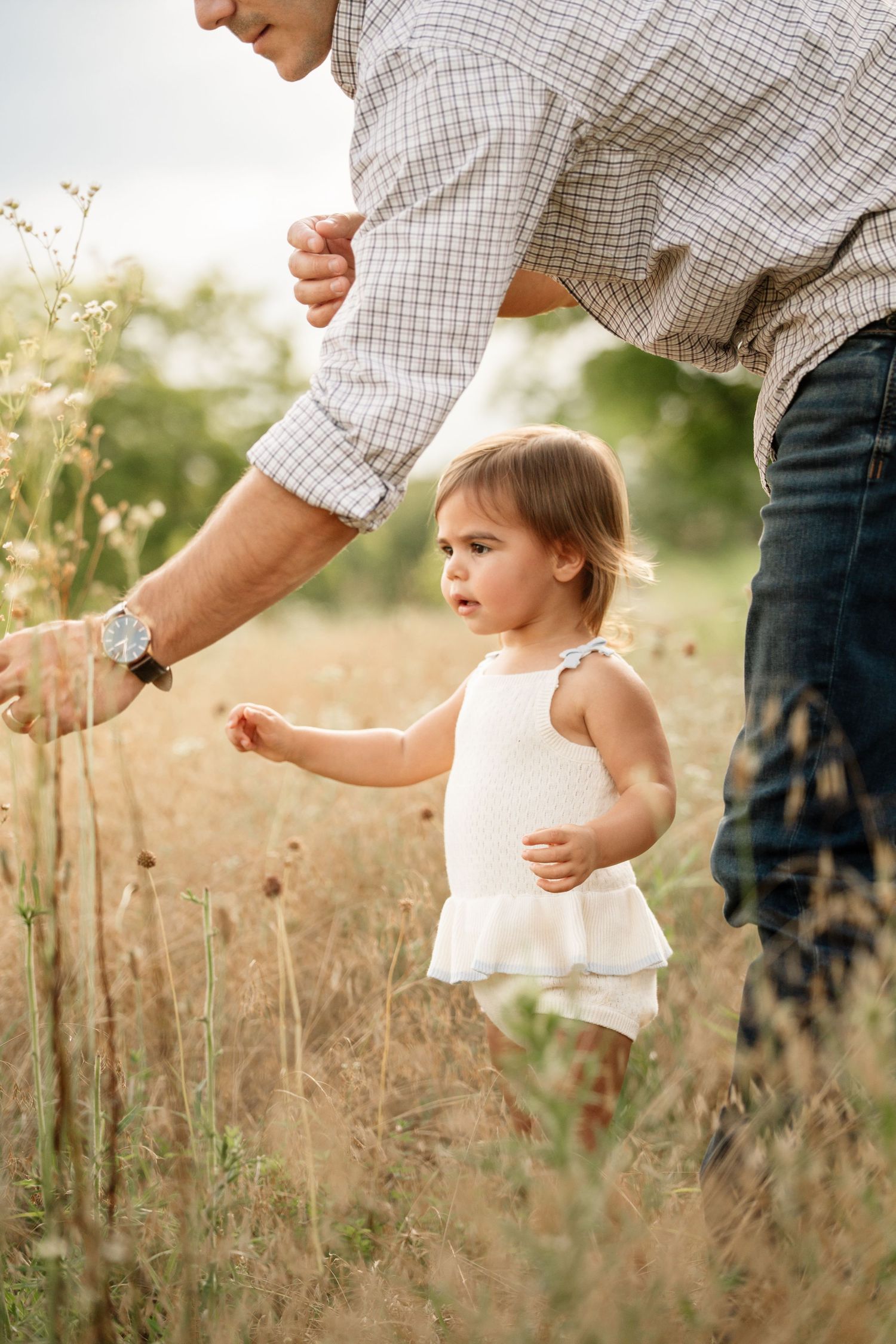 A toddler girl in white romper walks through tall grass alongside her father in a sunlit meadow.