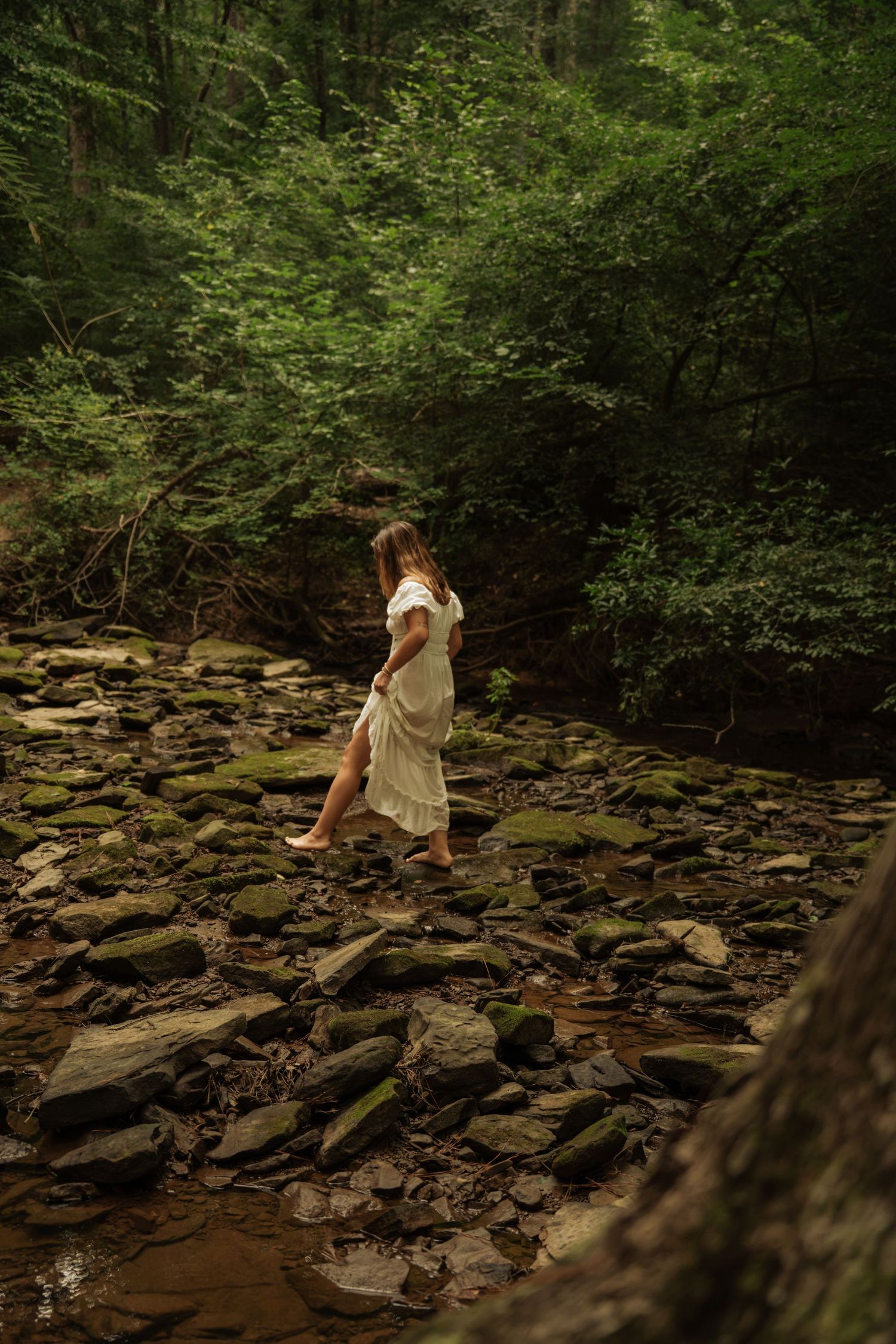 Barefoot woman in white off-shoulder dress walking across rocky creek bed surrounded by green forest.