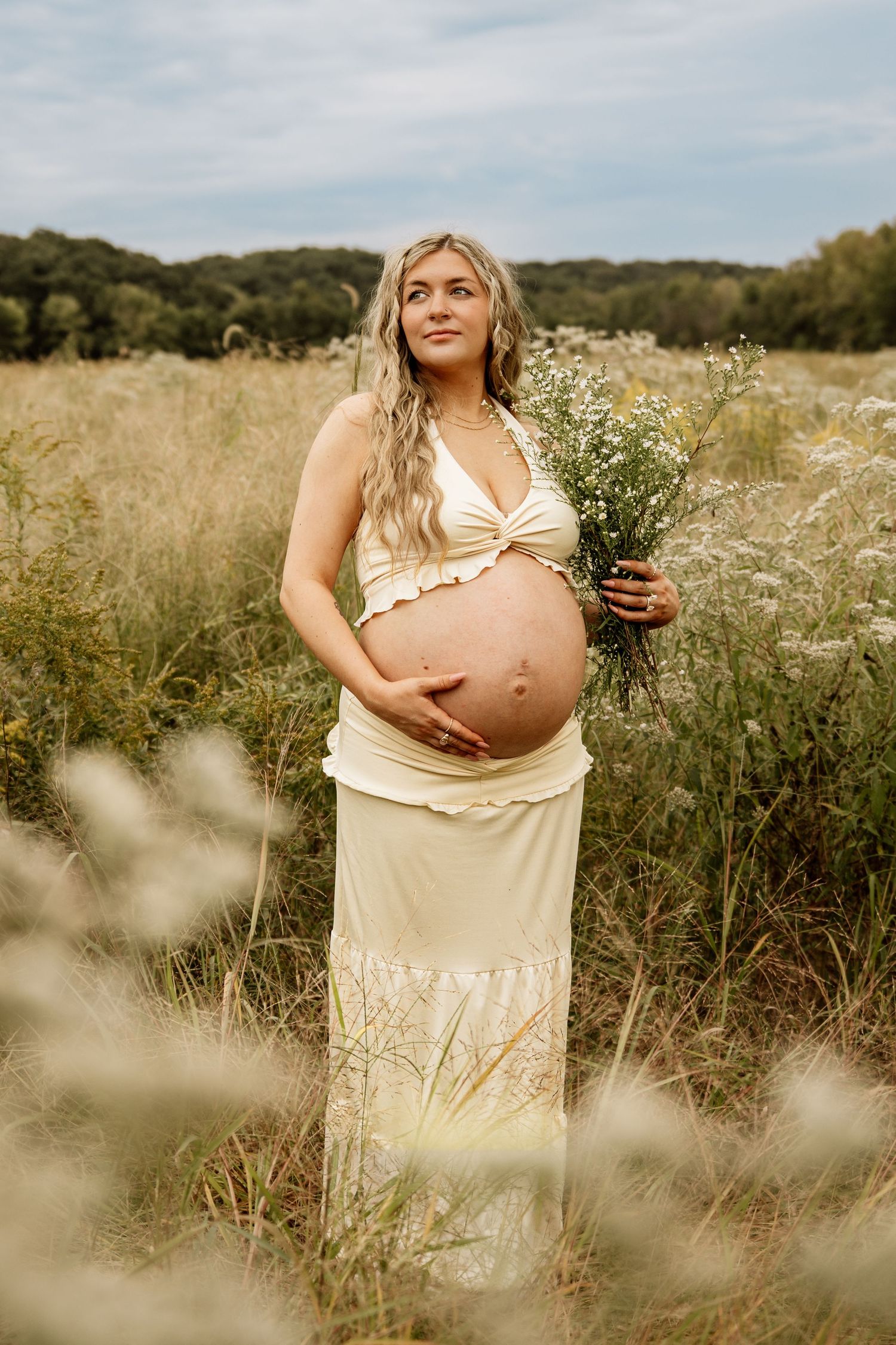 Pregnant woman in cream halter maternity dress holds wildflowers while standing in a meadow field.