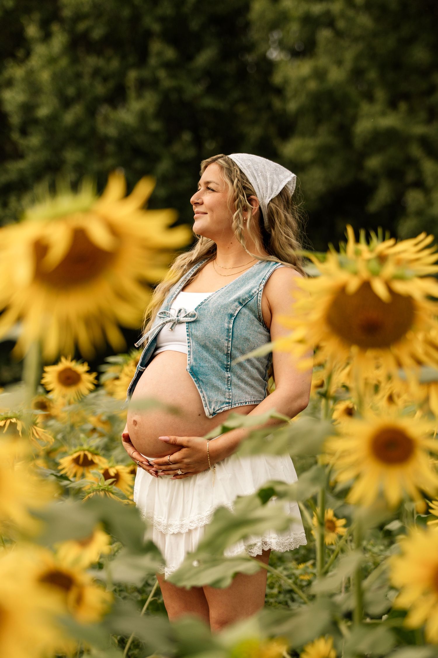 Pregnant woman in denim vest and white dress cradles baby bump among sunflowers in a lush green field.