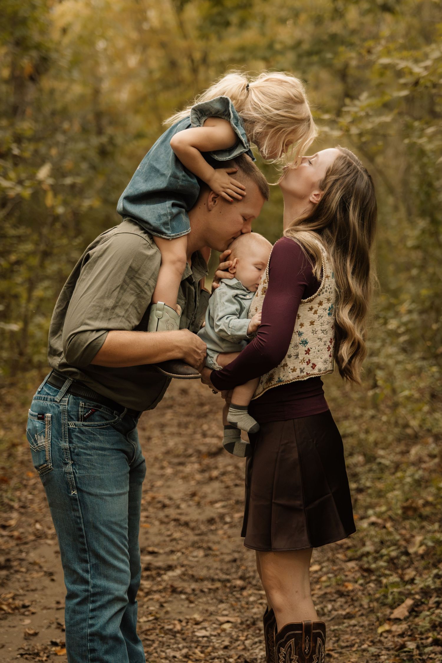 Young family sharing a sweet kiss on a woodland trail, father holding baby and toddler on shoulders.