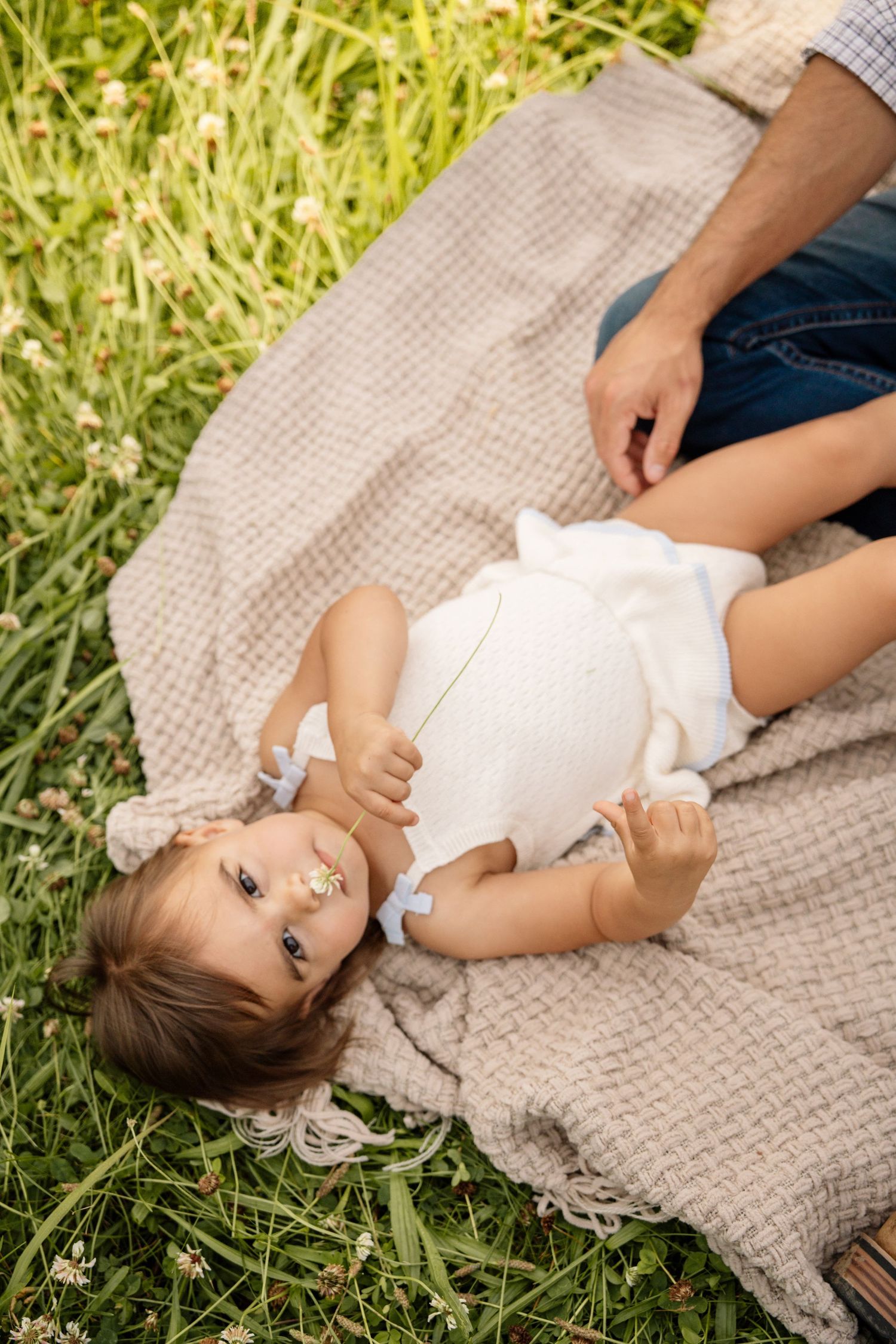 Adorable toddler girl lying on blanket in green grass field with parent nearby.
