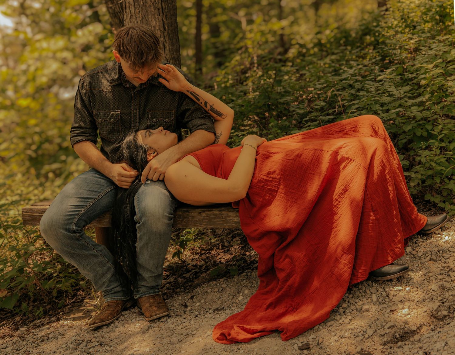 Couple in romantic outdoor maternity shoot, woman in red gown resting on mans lap on bench in forest.