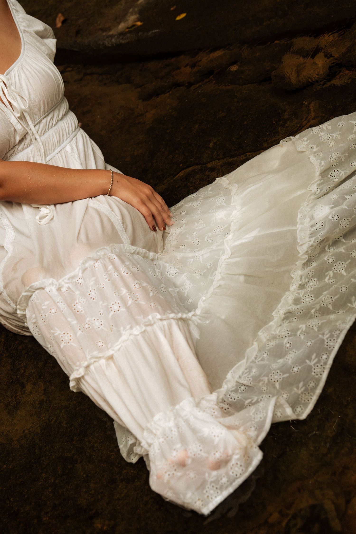 Close-up of a woman in a white lace dress sitting on rocks in a forest.