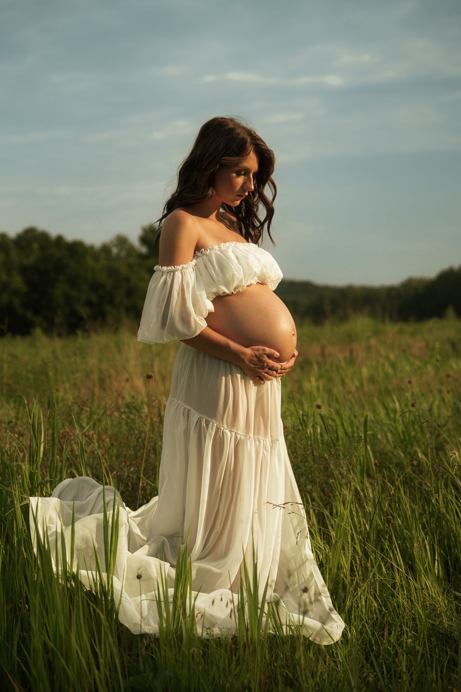 Beautiful pregnant woman in flowing white dress standing in a lush green meadow at sunset.
