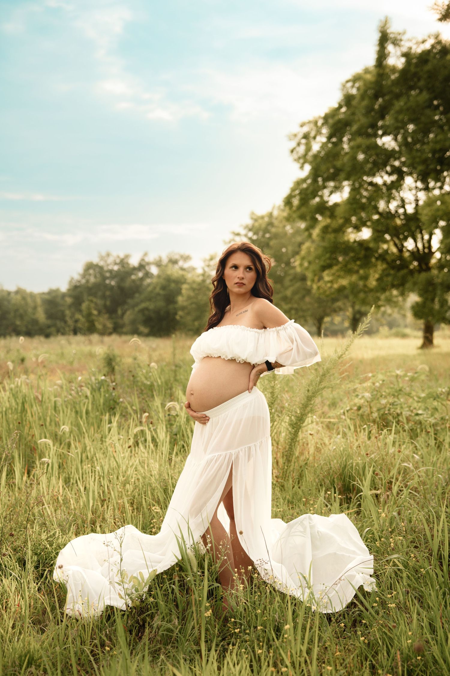 Pregnant woman in elegant white maternity dress poses in a lush green meadow under a blue sky.