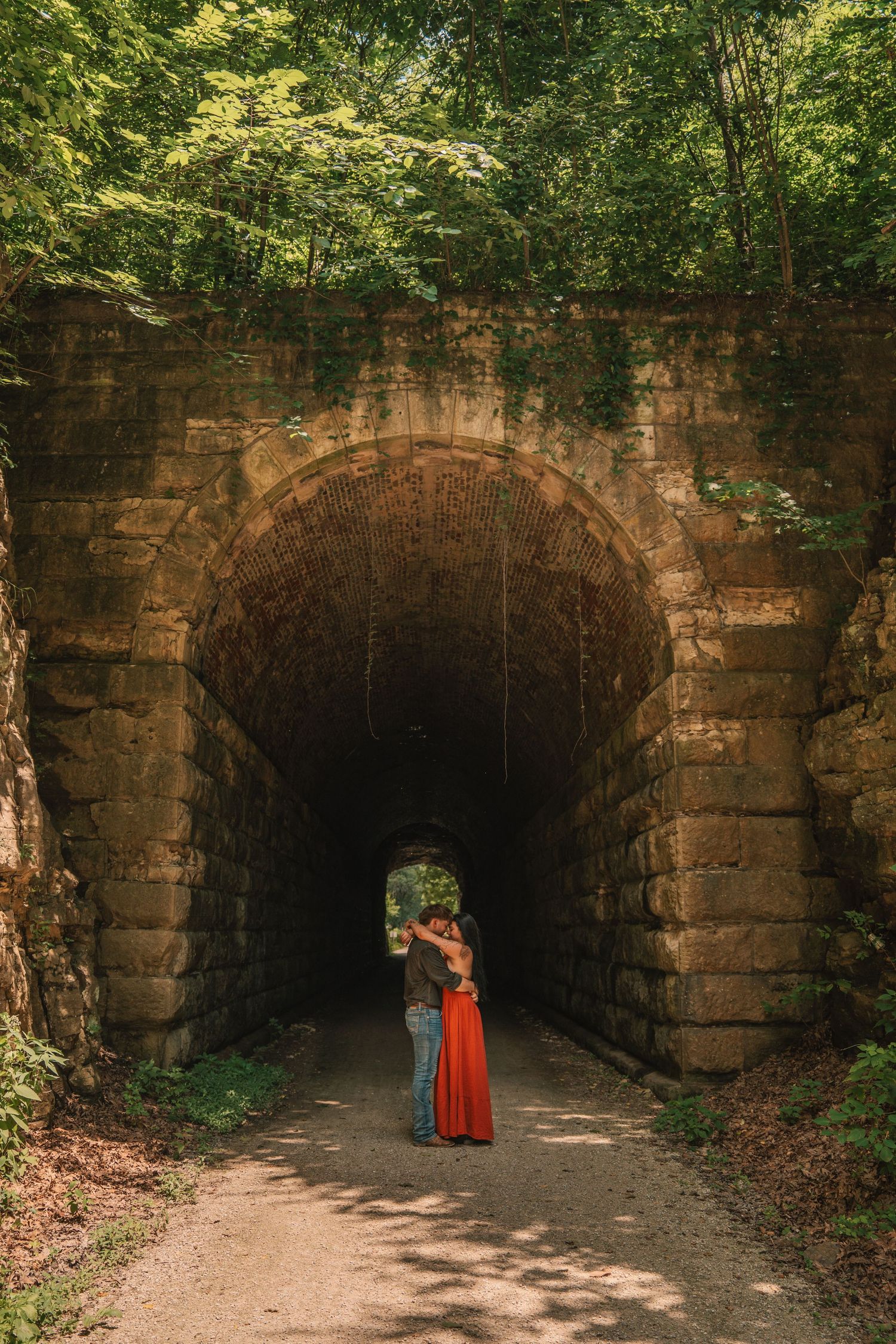 Couple embracing in a romantic kiss under a historic stone arch tunnel surrounded by lush green forest trees.