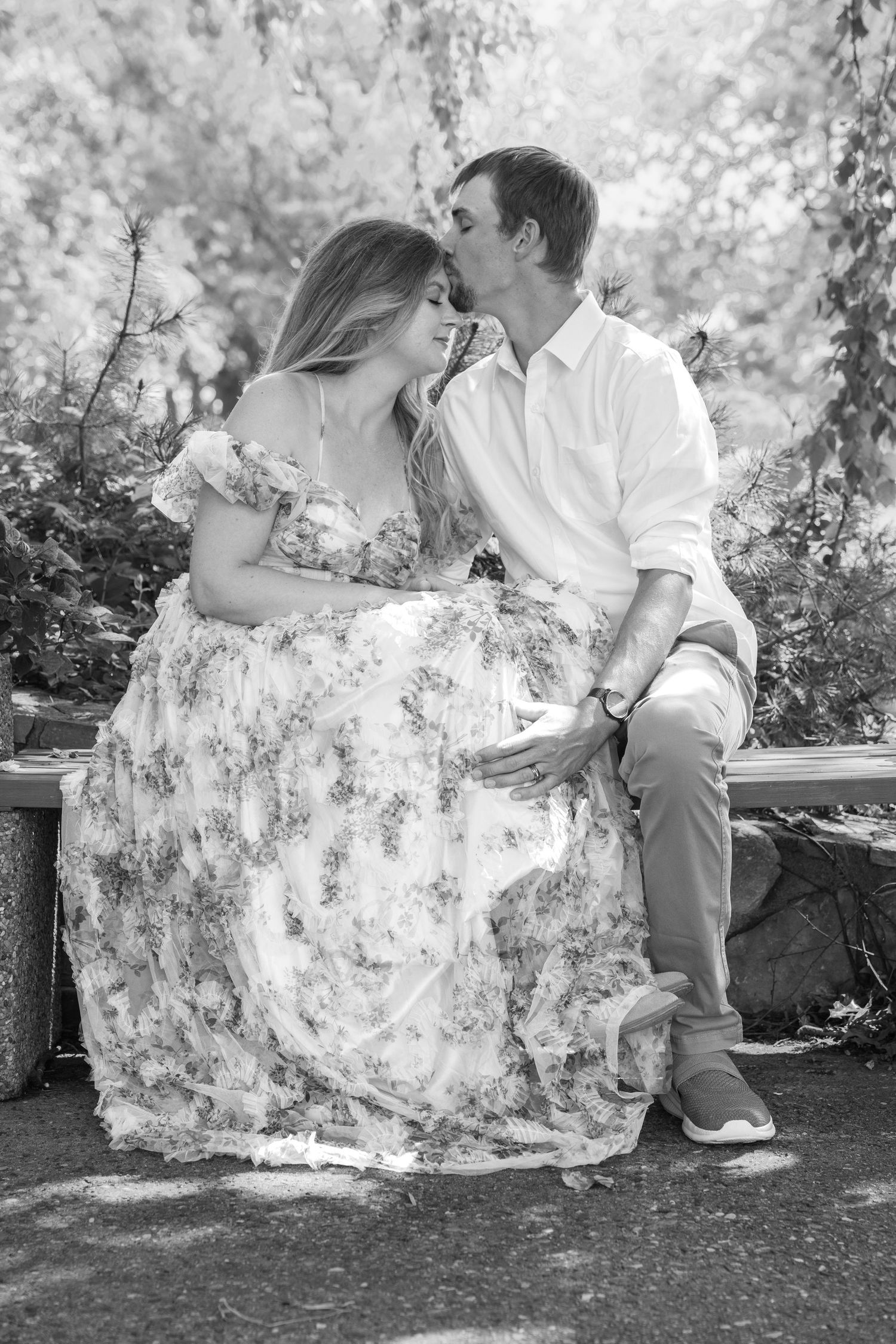 Black and white photo of a couple sitting outdoors, man kissing pregnant woman's forehead in a floral dress.