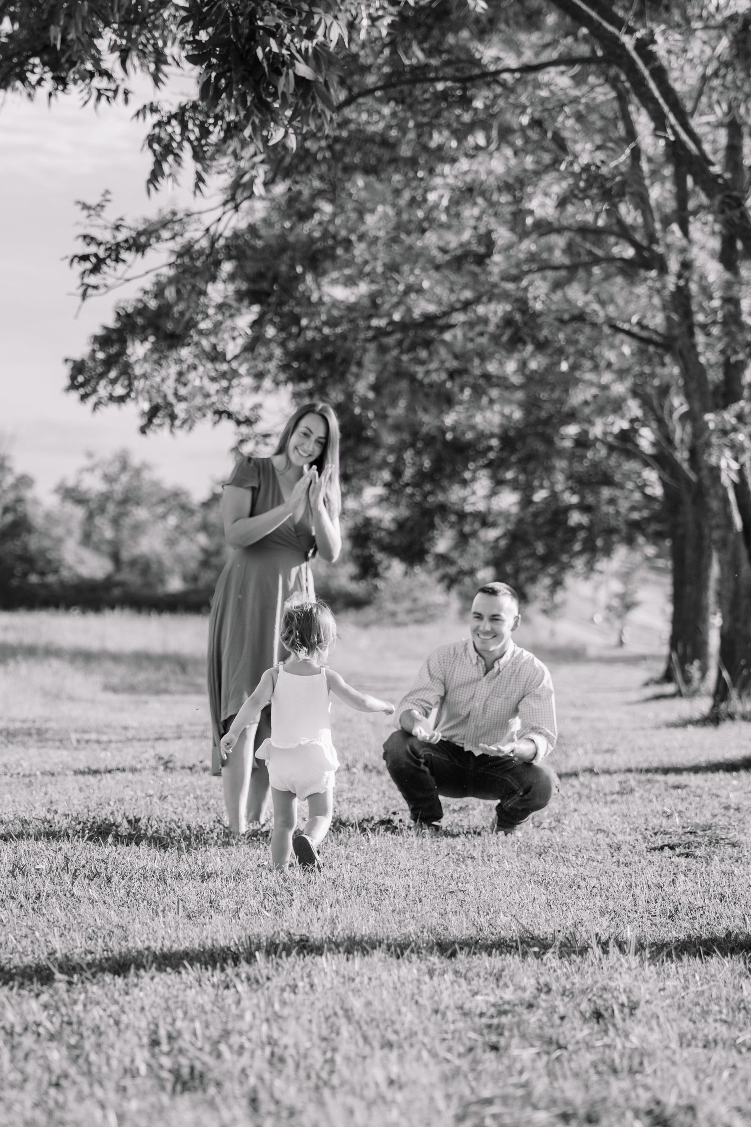 Black and white photo of a happy family with a toddler taking first steps outdoors in a park.
