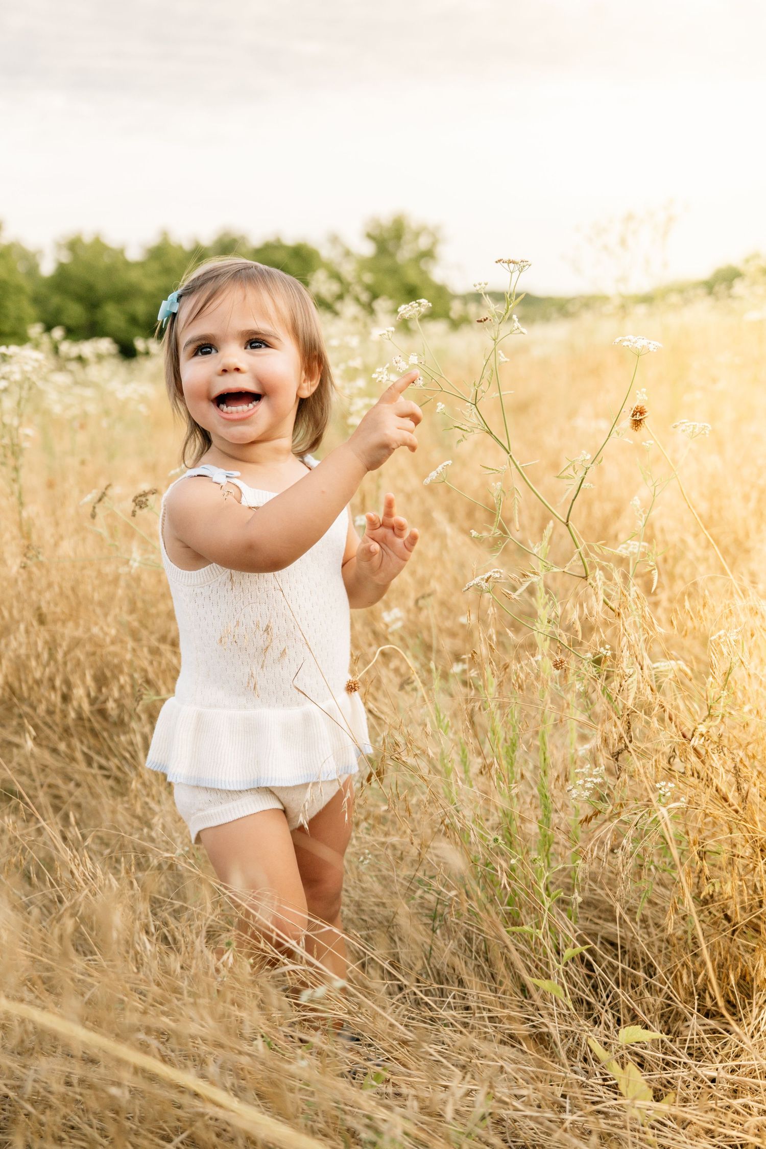 Cheerful toddler girl in white romper playing in golden meadow field at sunset.