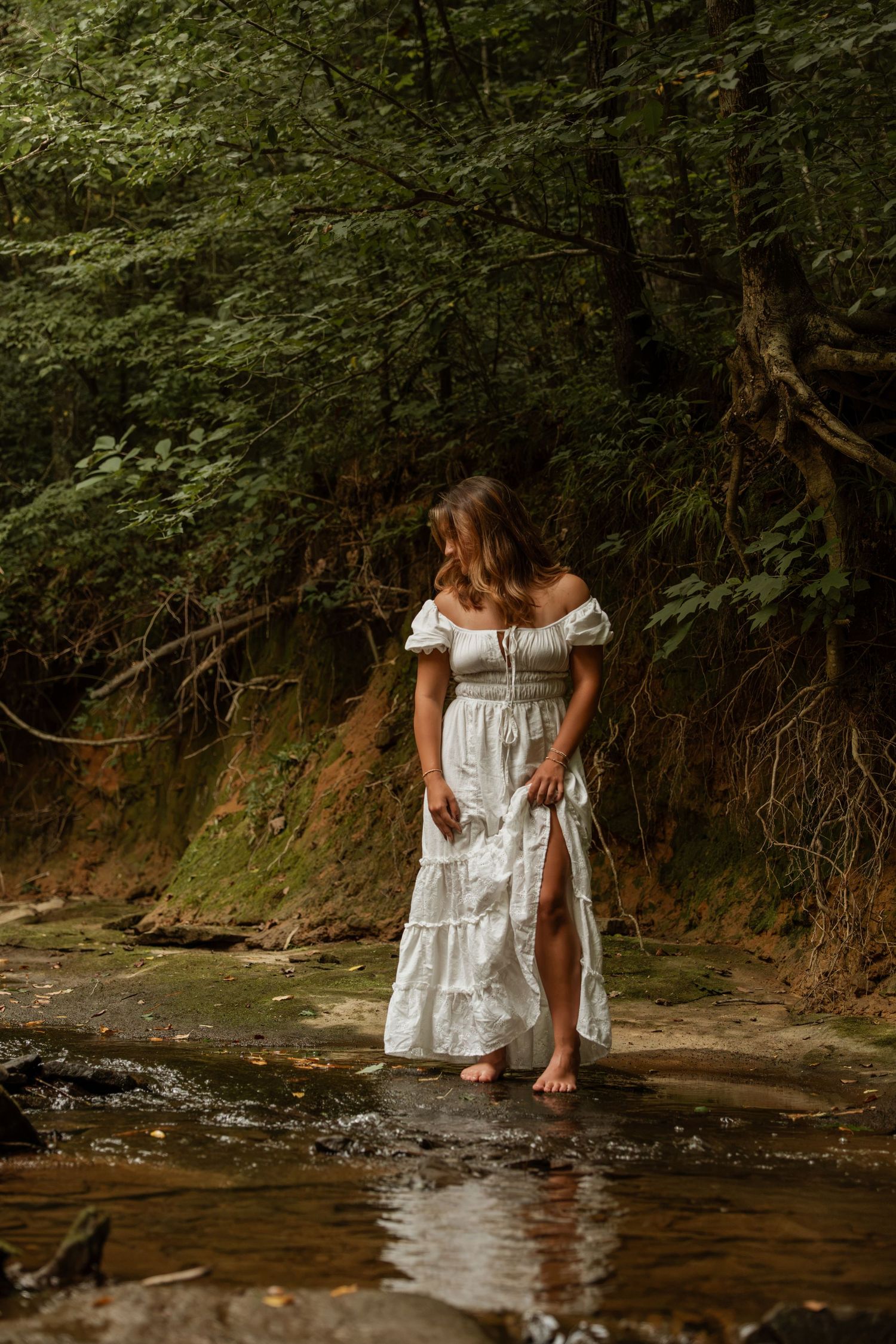 Woman in white tiered dress wading barefoot through a peaceful woodland stream.