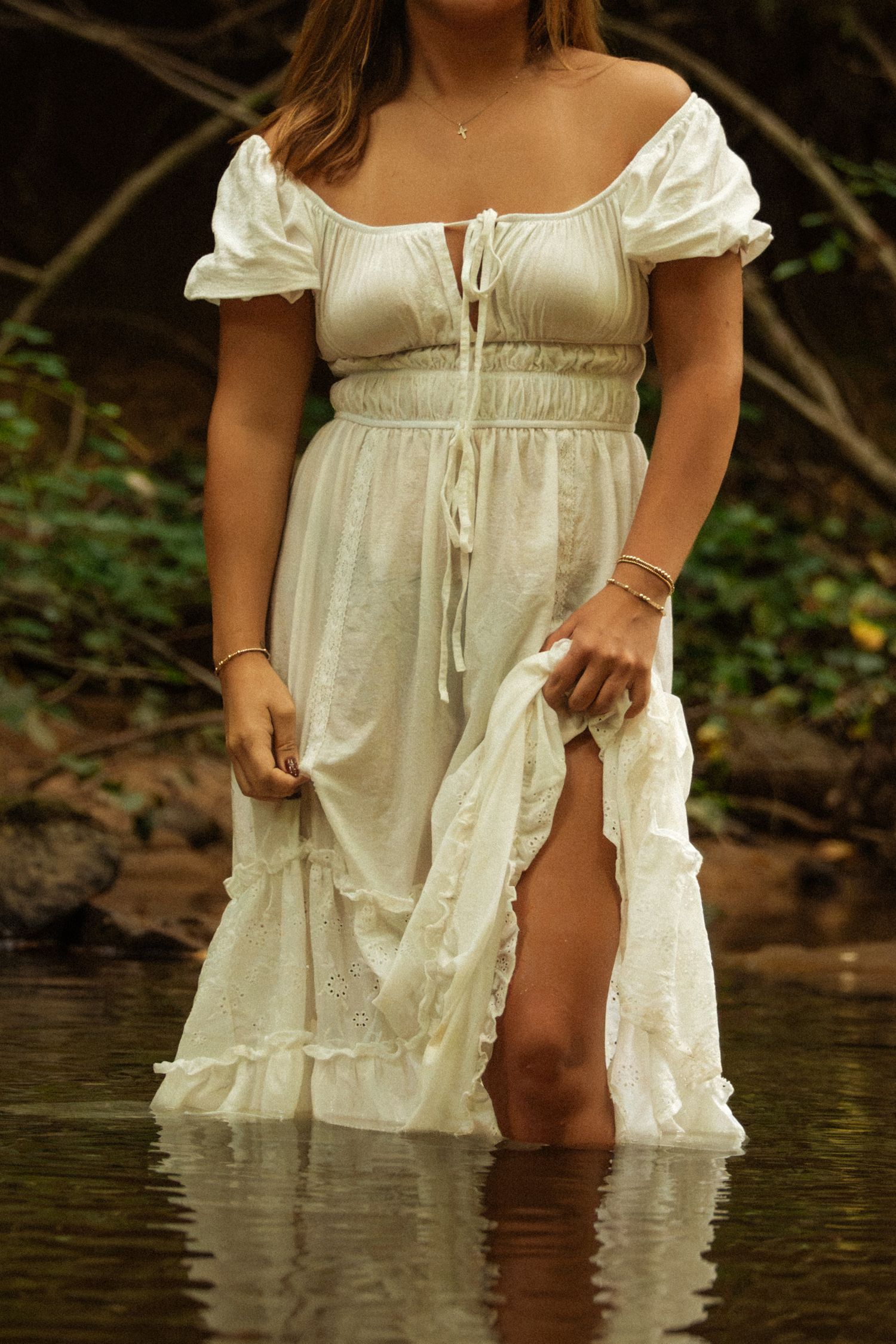 Woman in white off-shoulder ruffle maxi dress standing in shallow water with lush green forest background.