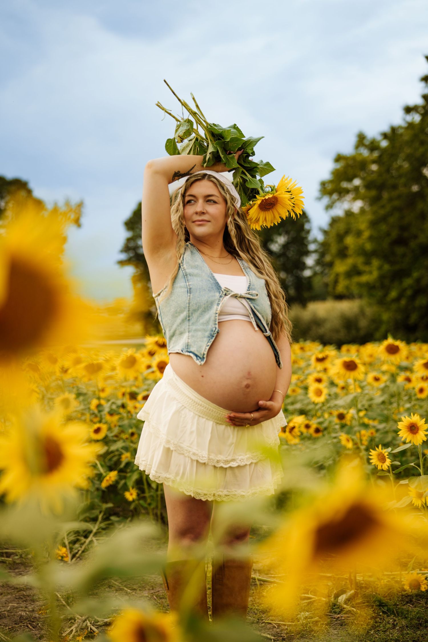 Pregnant woman in denim vest and white skirt holds sunflowers in a vibrant sunflower field.