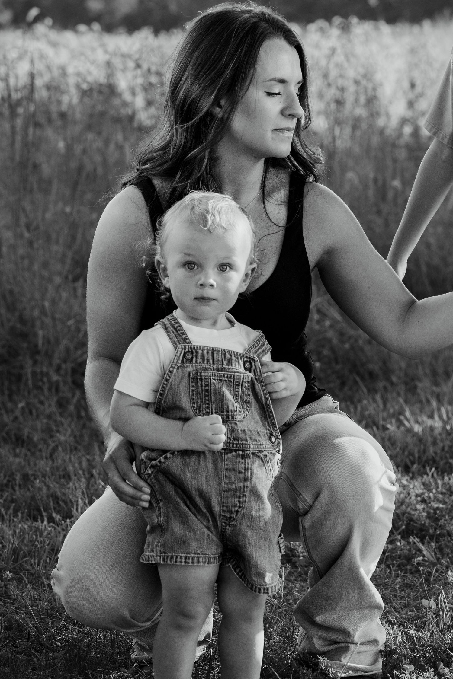 Black and white photo of a young mother kneeling in a field with her toddler in denim overalls.