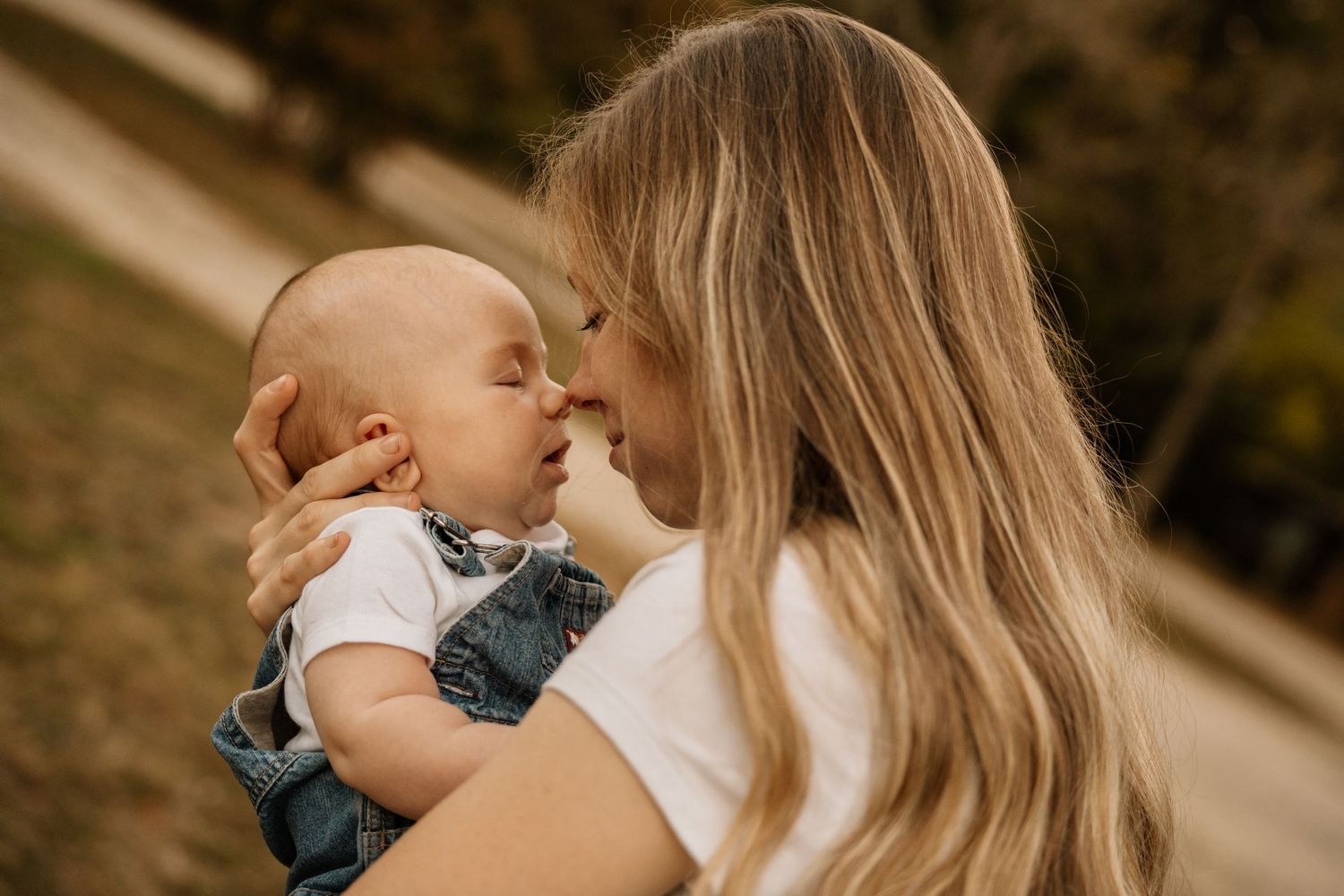 Loving mother with blonde hair touching noses with her baby boy outdoors in warm natural light.