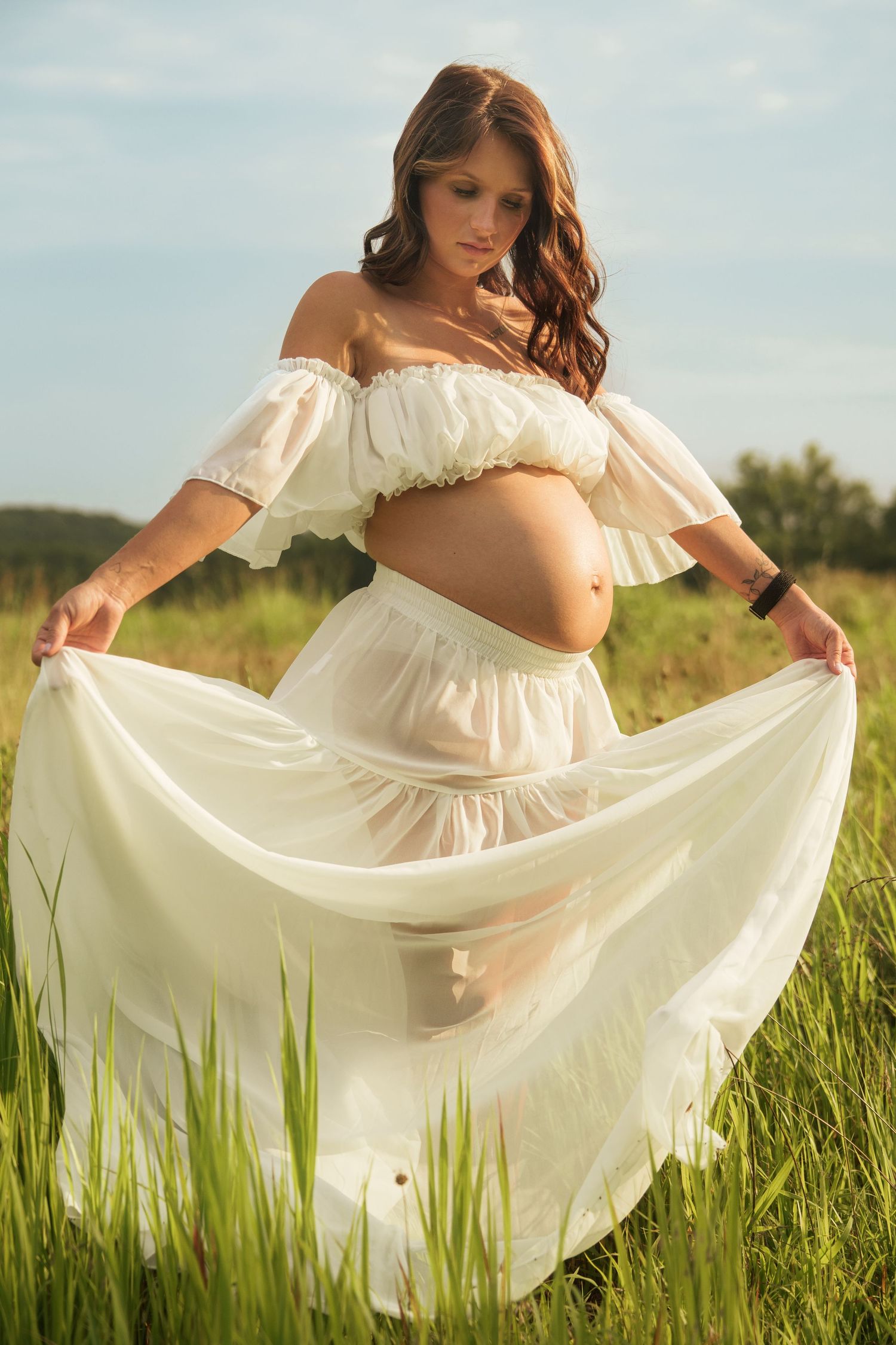 Pregnant woman in white off-shoulder dress spreading her skirt in a bright summer meadow.