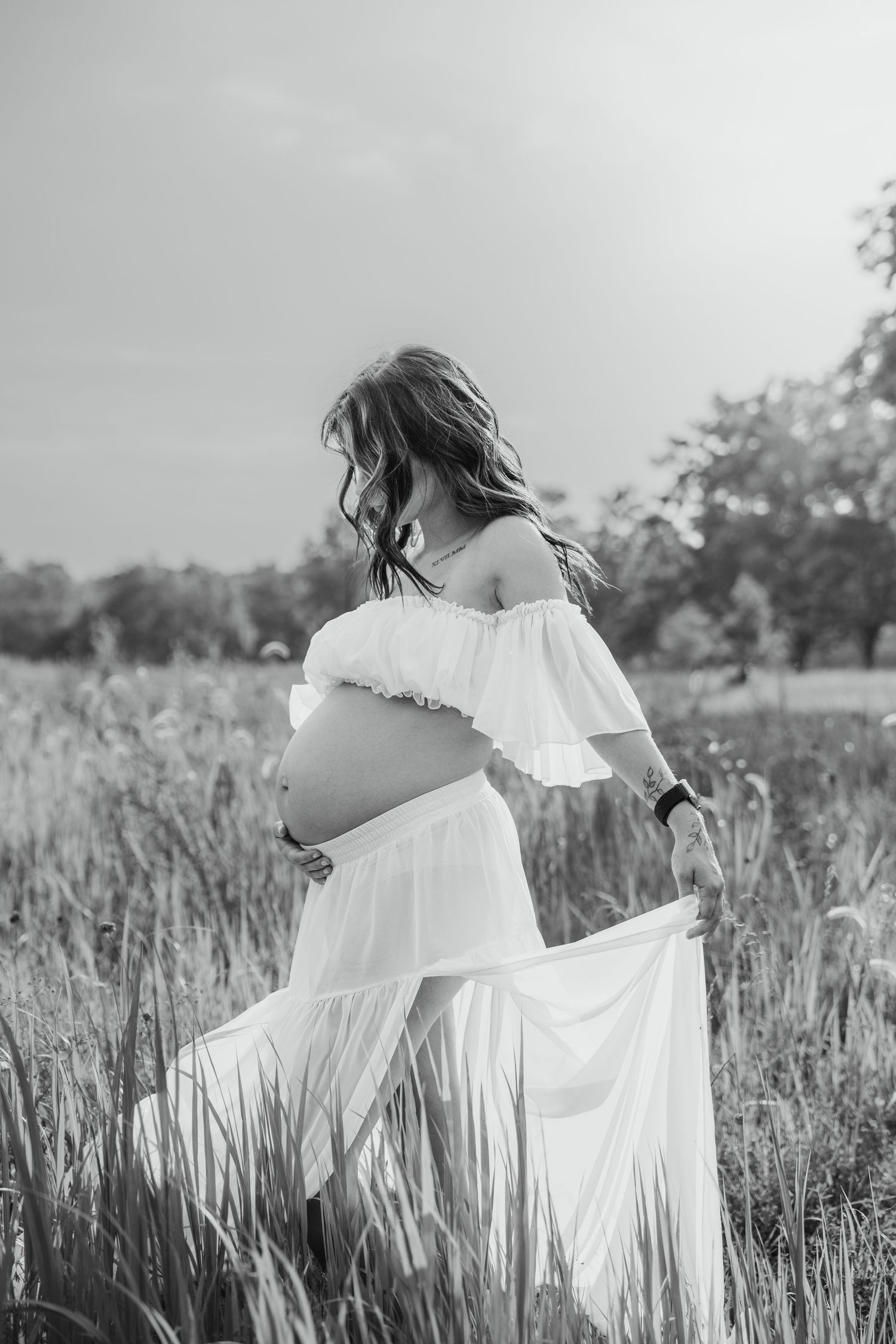 Black and white photo of pregnant woman in white off-shoulder dress standing in a field, cradling her baby bump.