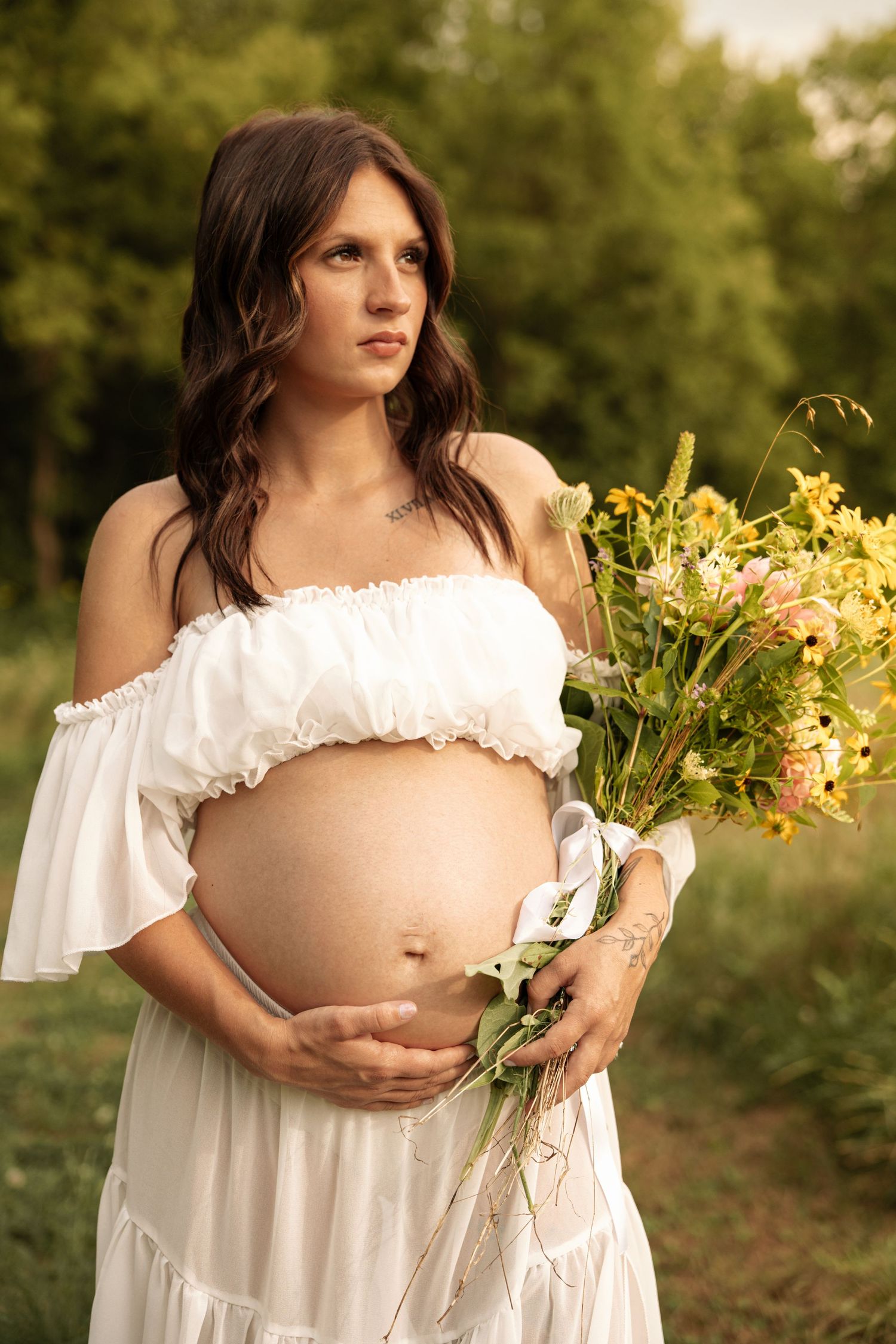Pregnant woman in white off-shoulder dress holds wildflower bouquet outdoors, cradling her baby bump in a lush green setting.