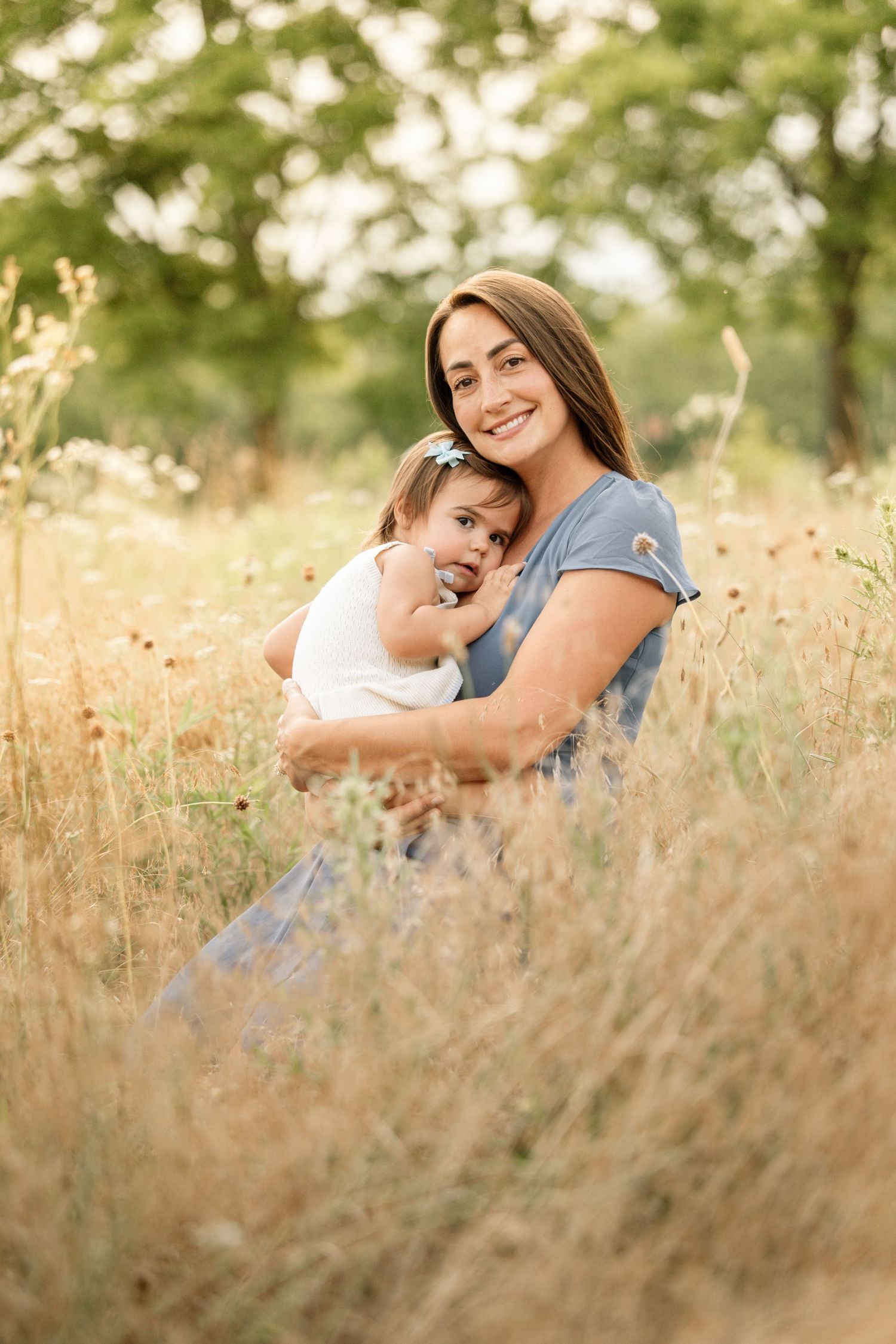 Smiling mother hugging happy toddler girl sitting in golden wildflower meadow outdoors.