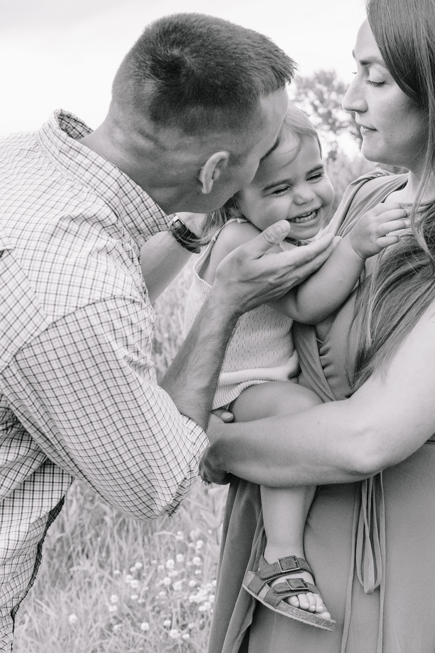 Black and white photo of parents lovingly kissing their laughing toddler daughter outdoors.