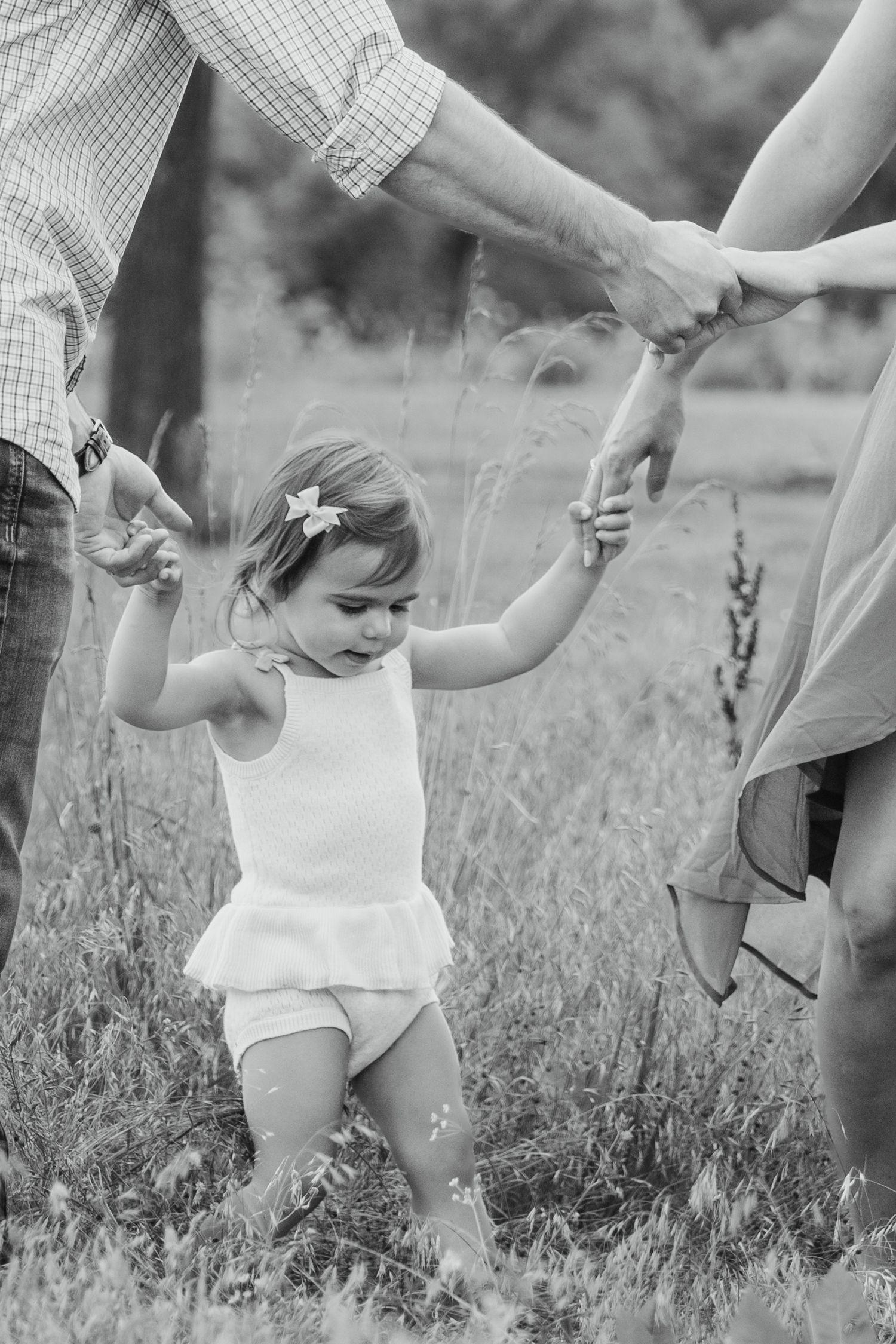 Black and white photo of a toddler girl holding parents hands while learning to walk in a grassy field.